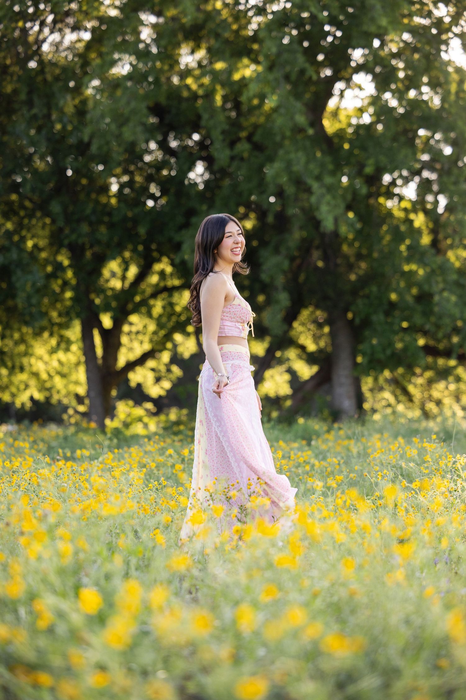 Smiling young woman in pink crop top and skirt standing in yellow wildflower field with green trees behind her.