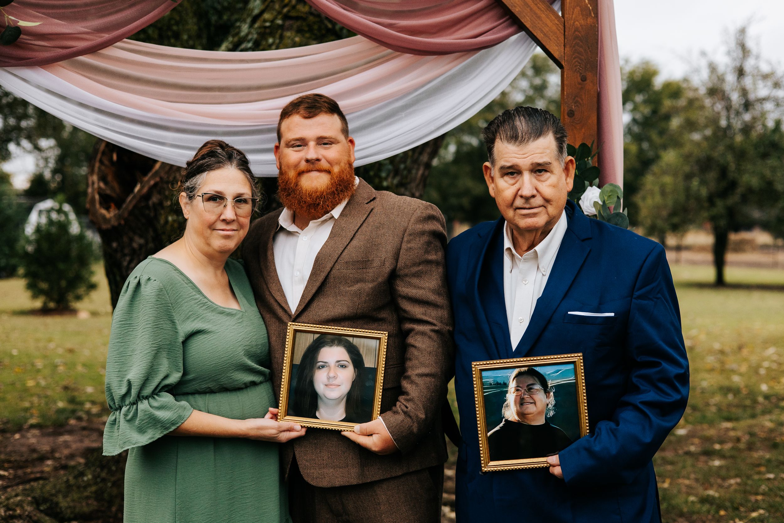 Three people holding framed photos at an outdoor wedding ceremony, standing under a floral arch on a green lawn.