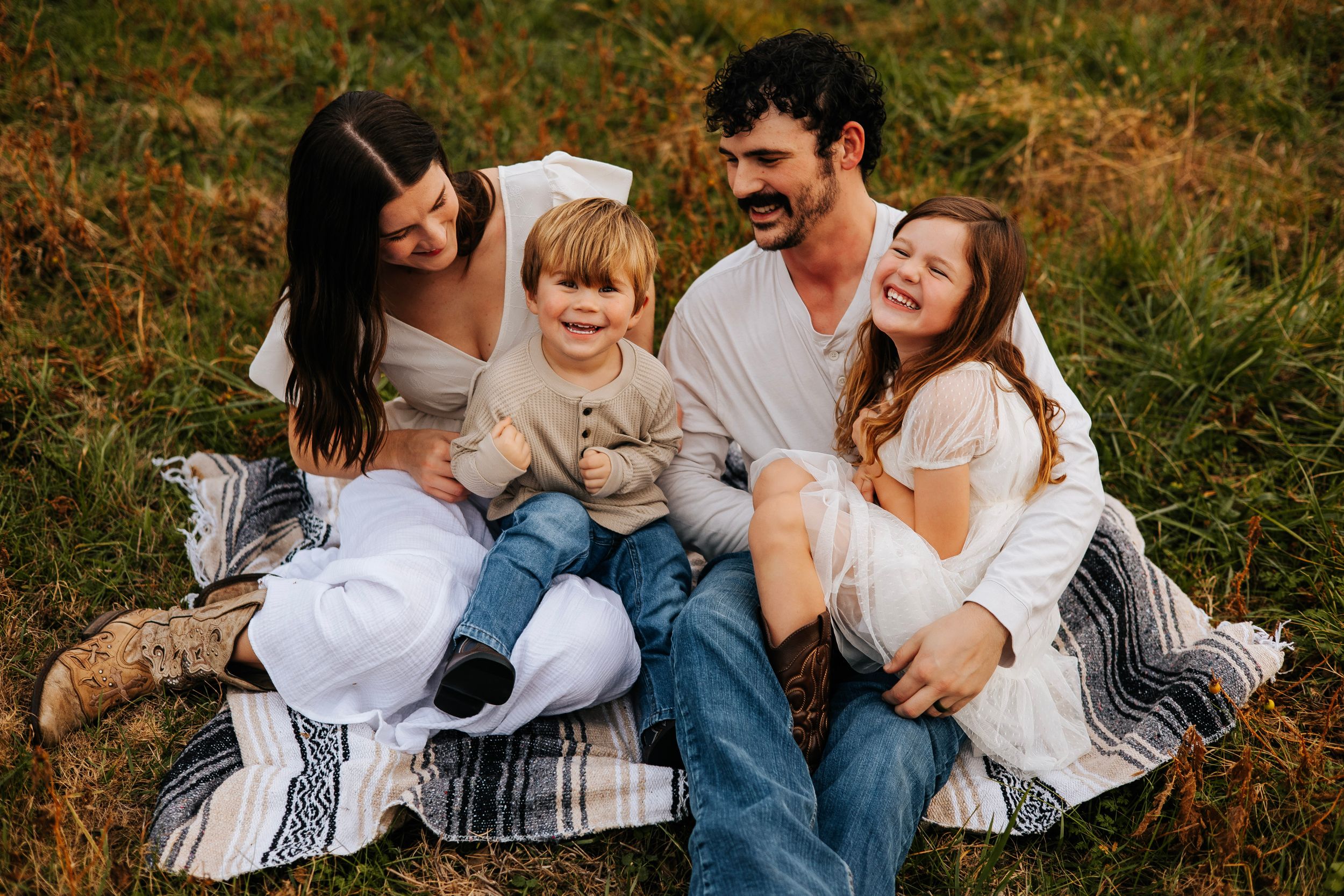 Happy family of four sitting on a blanket in a grassy field, laughing together outdoors.