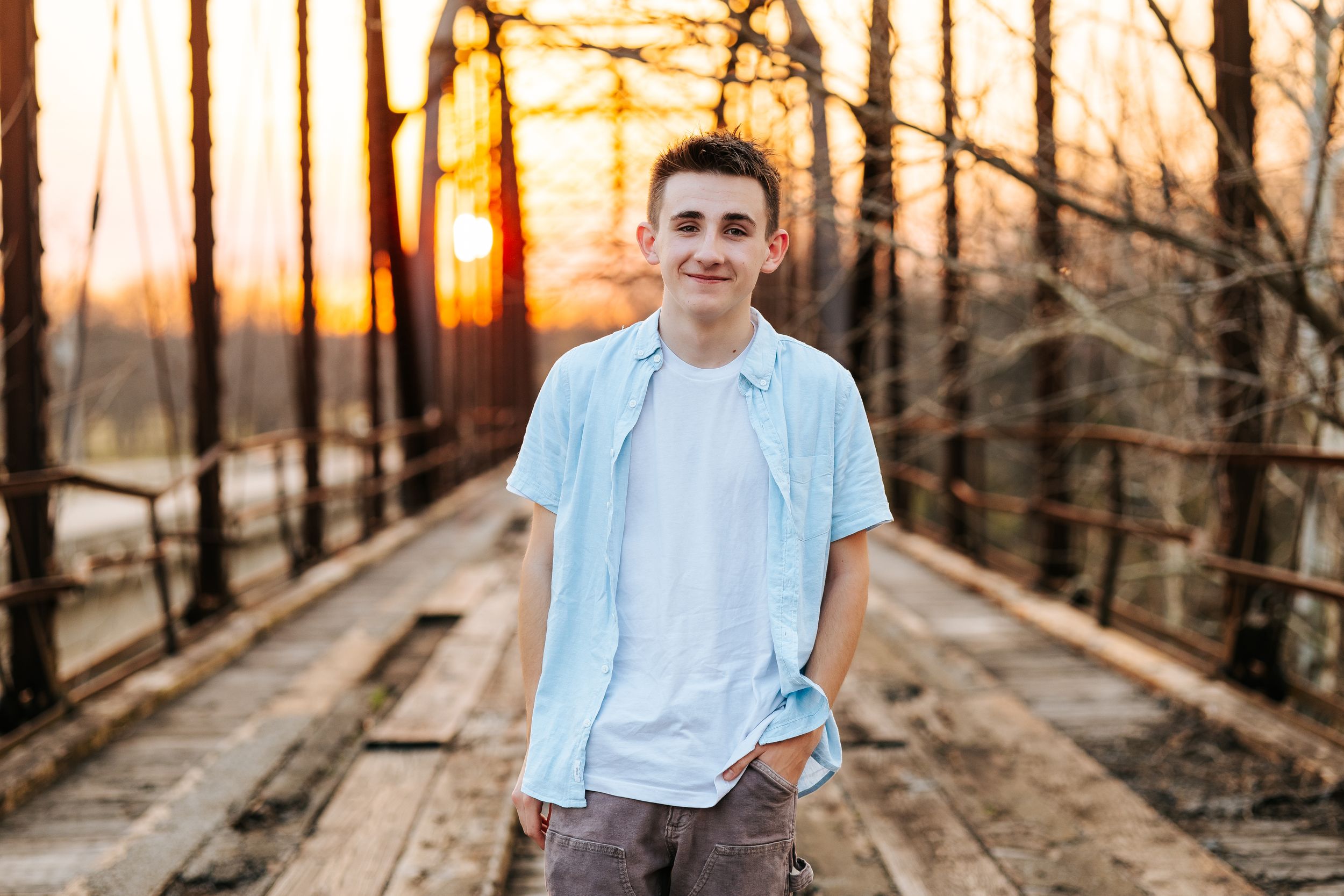 Teen boy in light blue shirt smiling on wooden bridge at golden sunset hour.