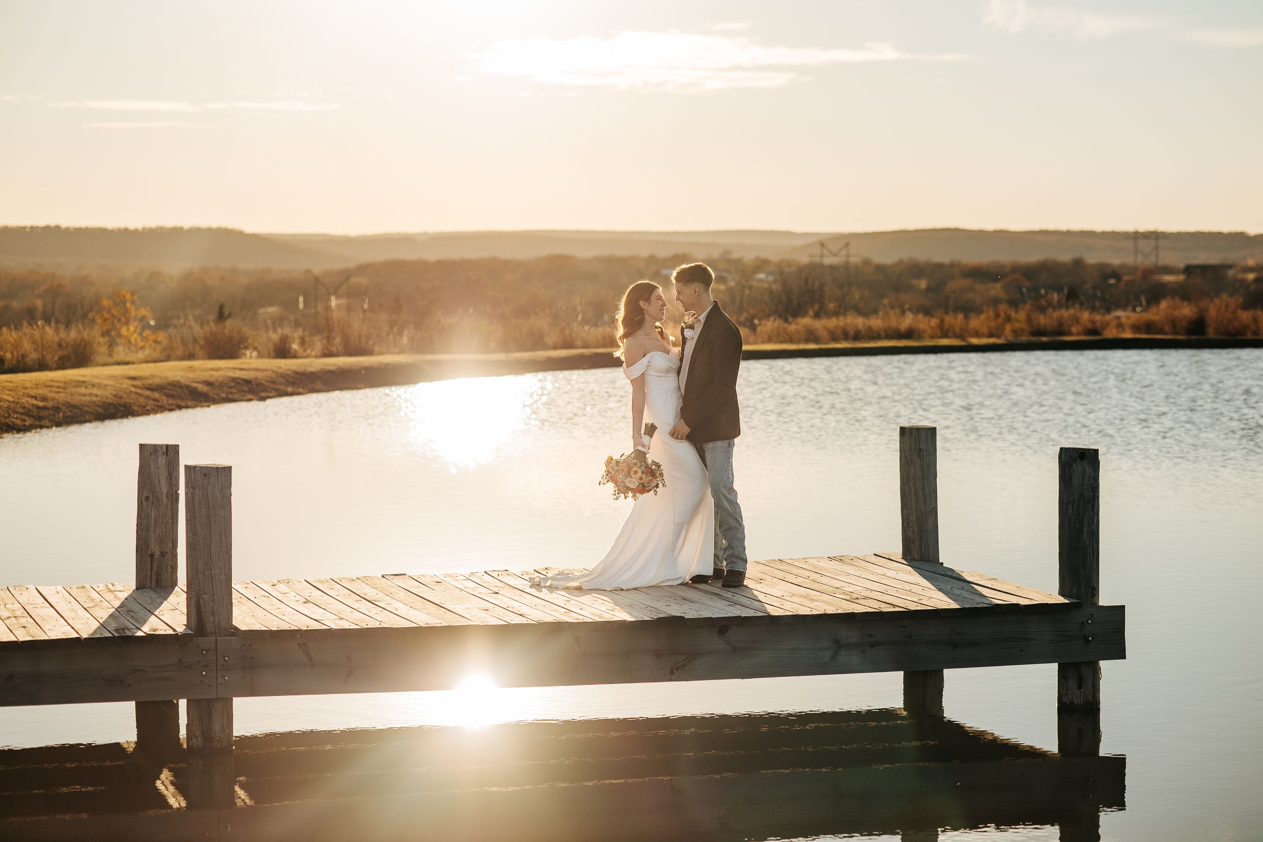 Bride and groom kissing on a wooden dock at sunset by a serene lake with golden light.