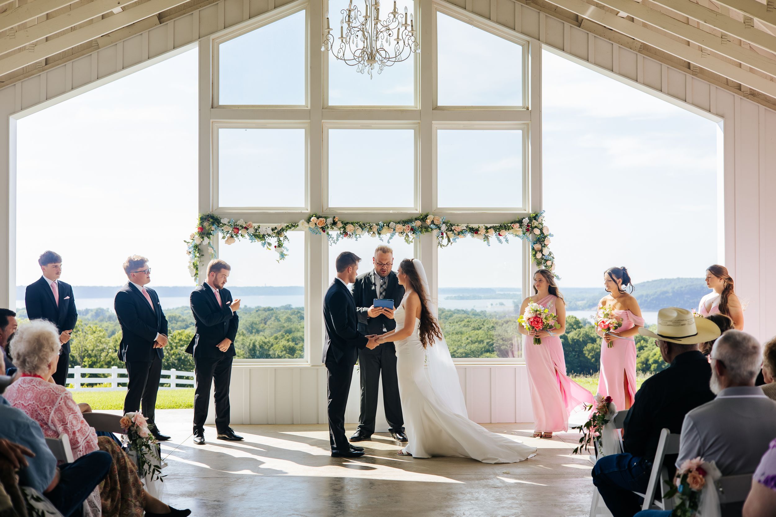 Elegant barn wedding ceremony with couple exchanging vows before large windows overlooking scenic countryside views.
