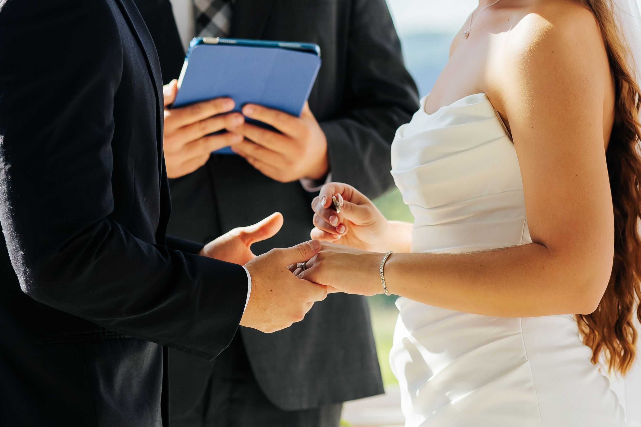 Bride placing a wedding ring on groom's finger during ceremony while officiant holds a tablet.