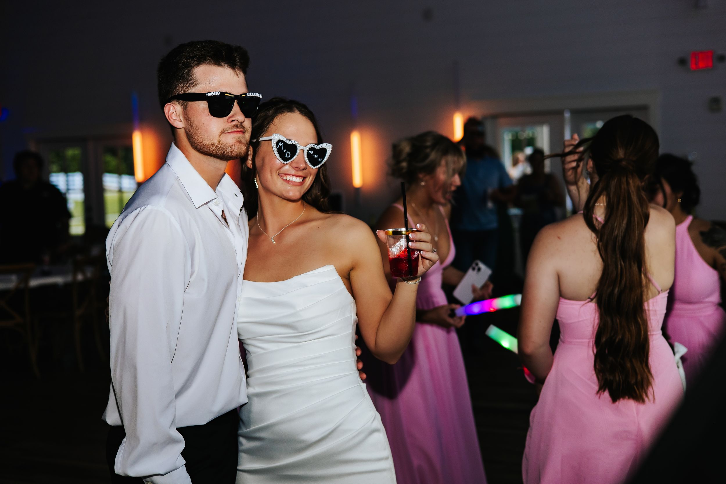 Bride and groom wearing fun sunglasses dancing together at their wedding reception with guests in background.