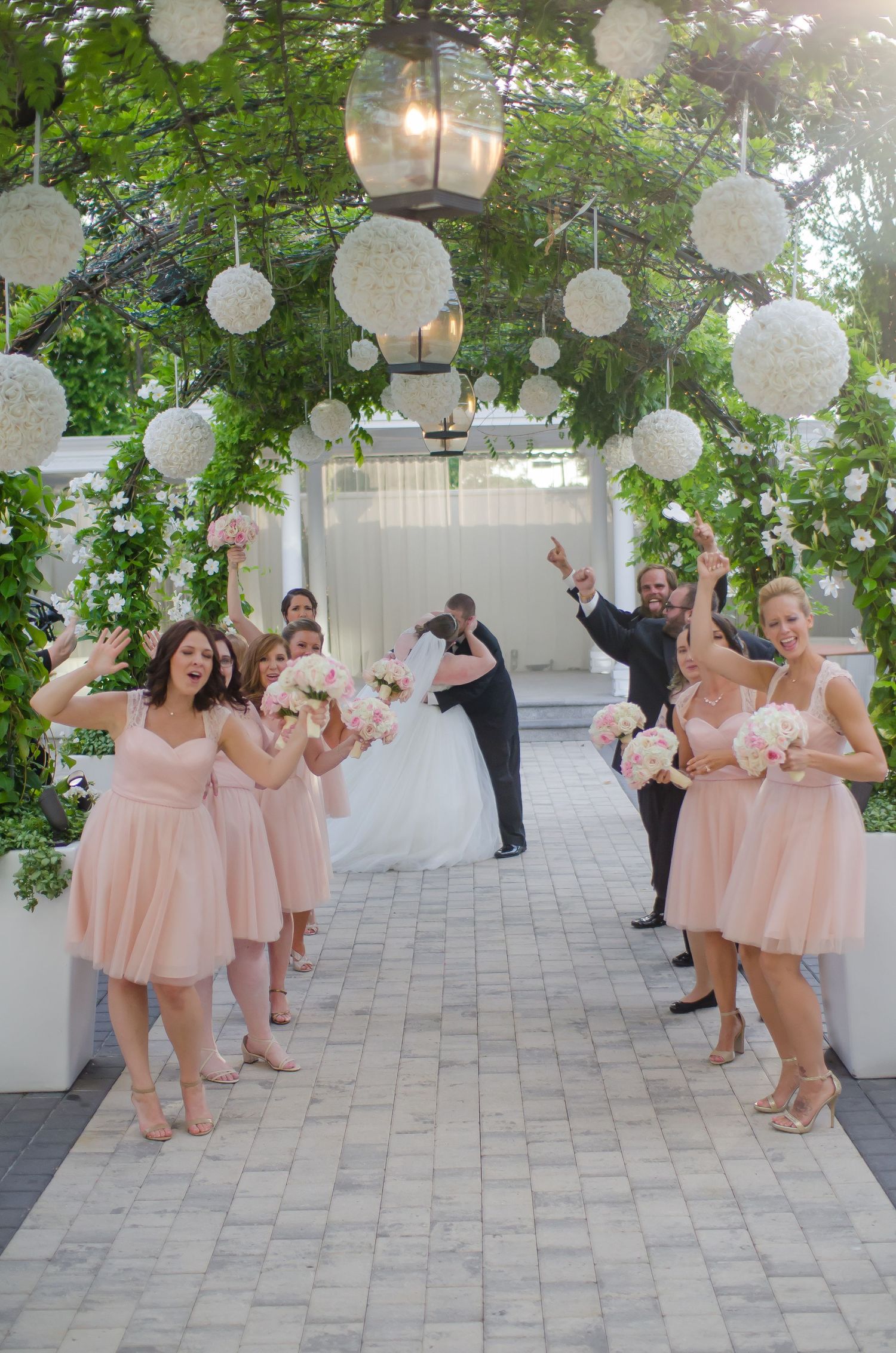 Bride and groom kiss under floral arch with bridesmaids in pink dresses celebrating at outdoor wedding ceremony.