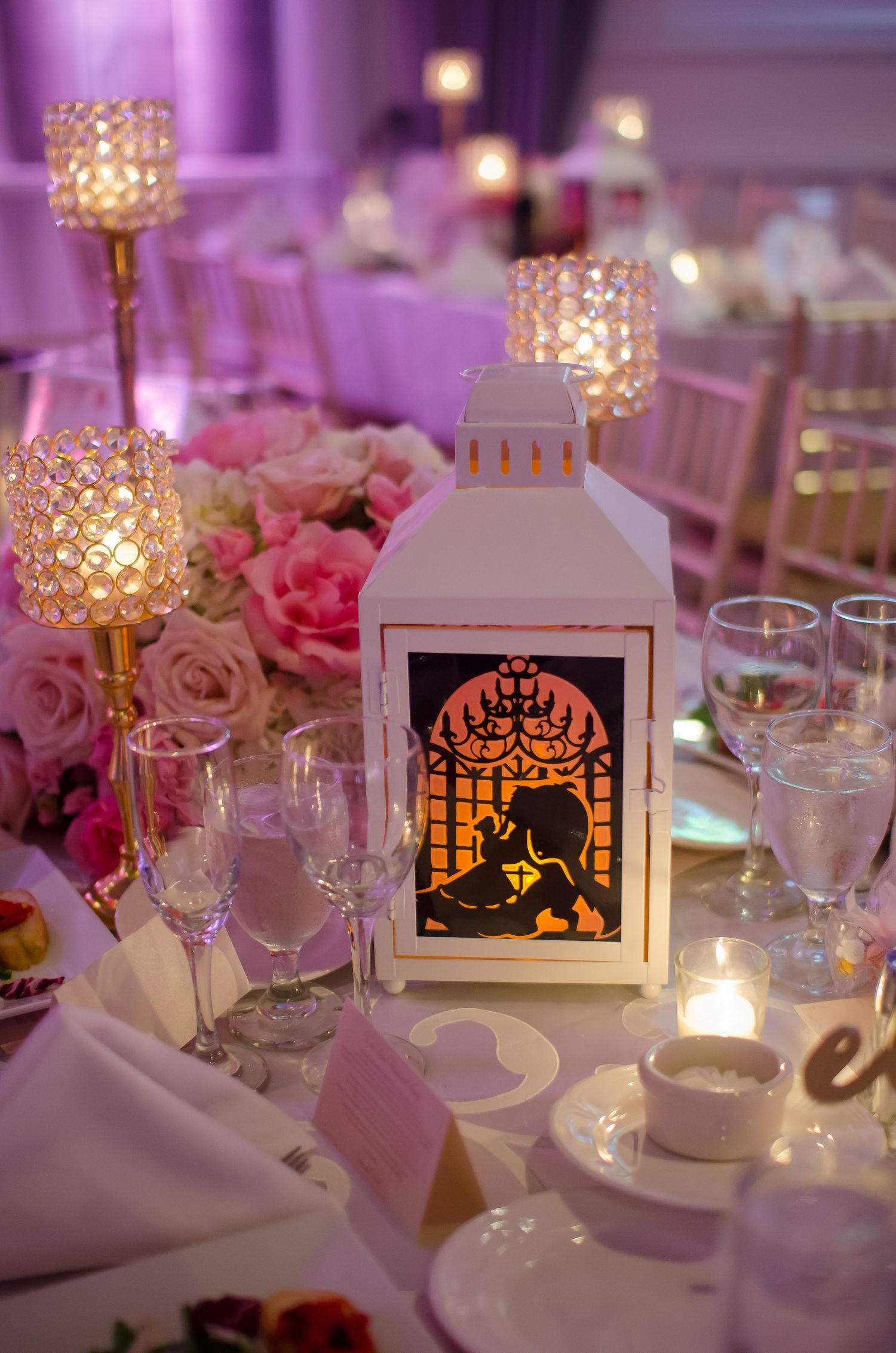 Elegant wedding reception centerpiece with white Beauty and the Beast lantern, crystal candle holders, and pink roses on a decorated table.