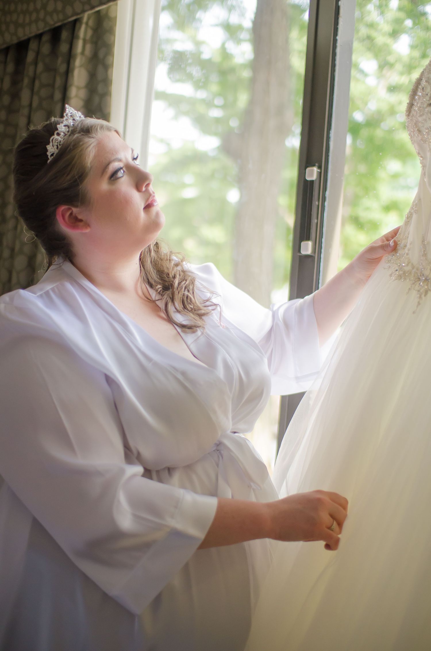 Bride in white robe and tiara admiring her wedding dress hanging by a sunlit window.