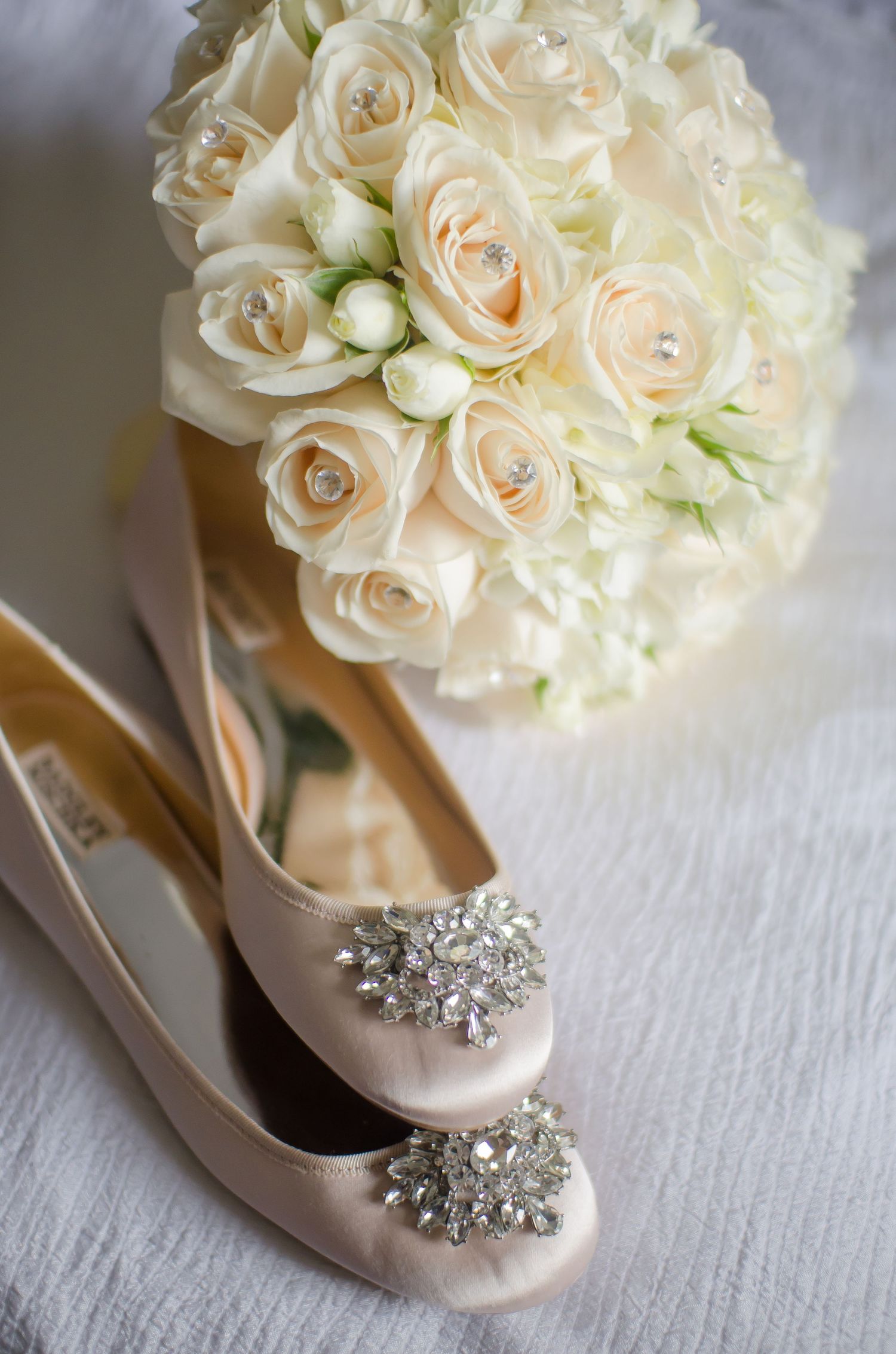 Elegant bridal flat shoes with crystal brooches beside a white and cream rose bouquet with pearl accents.
