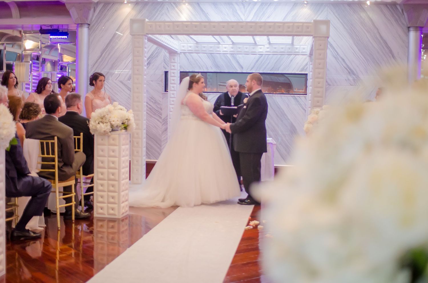 Bride and groom exchange vows at elegant indoor wedding ceremony with white floral arch and ivory aisle runner.