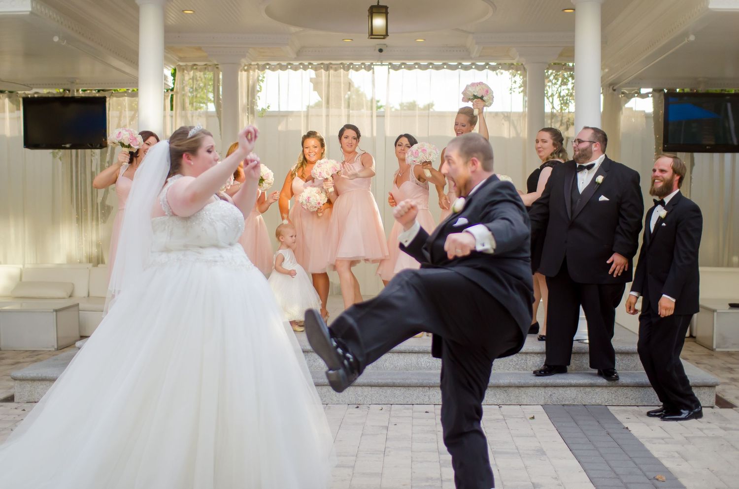 Bride and groom share a fun dance moment at wedding reception with bridesmaids and groomsmen cheering behind them.