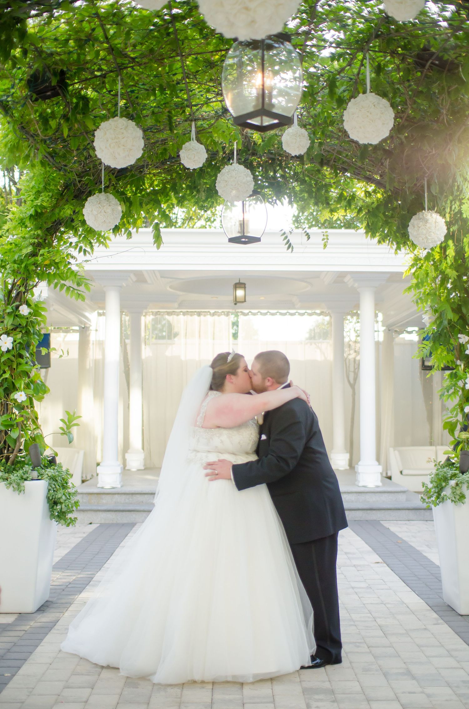 Bride and groom sharing first kiss under floral arch with white gazebo backdrop at outdoor wedding ceremony.