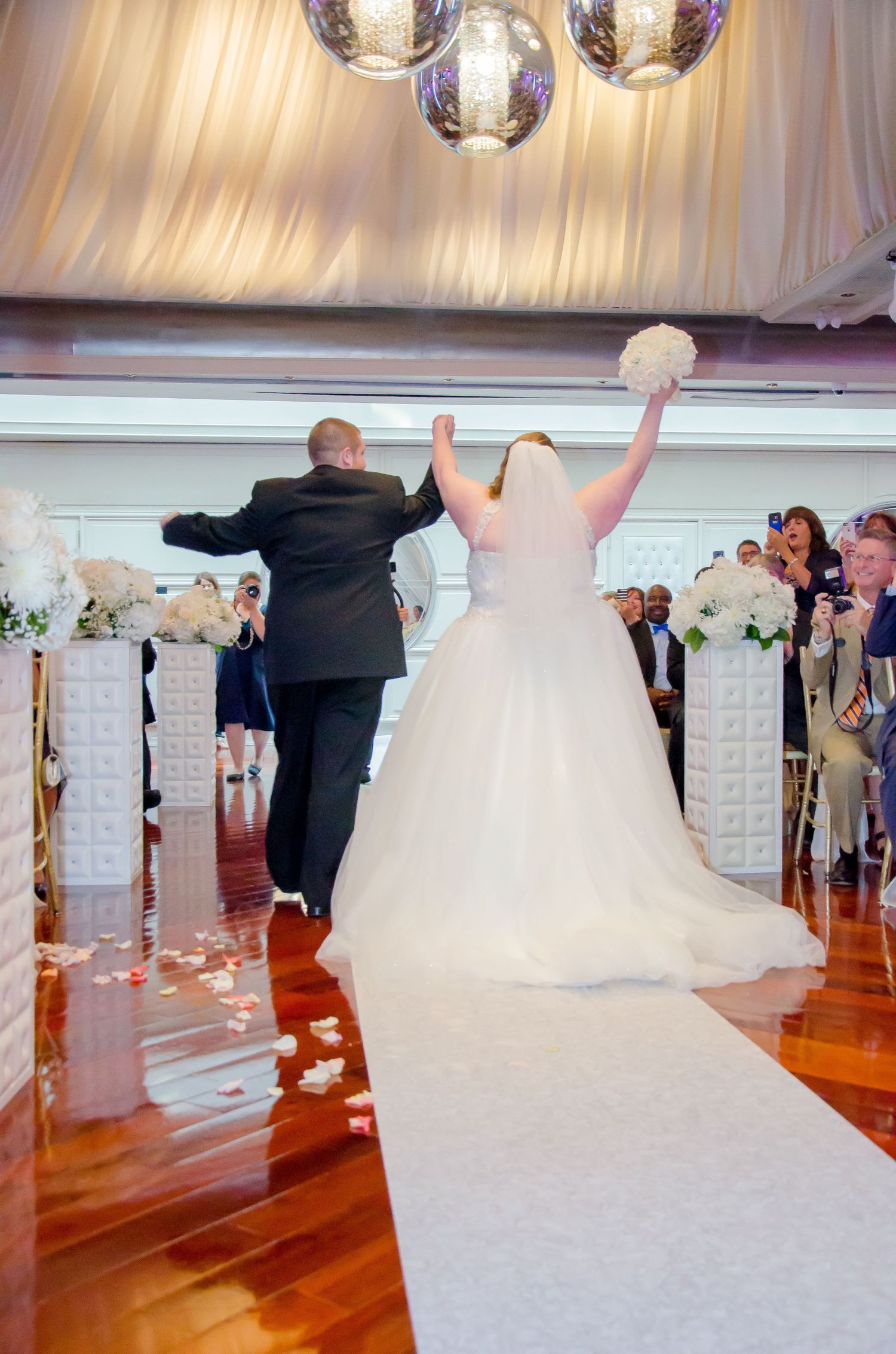 Bride and groom raising arms in celebration while walking down the aisle at elegant indoor wedding ceremony.
