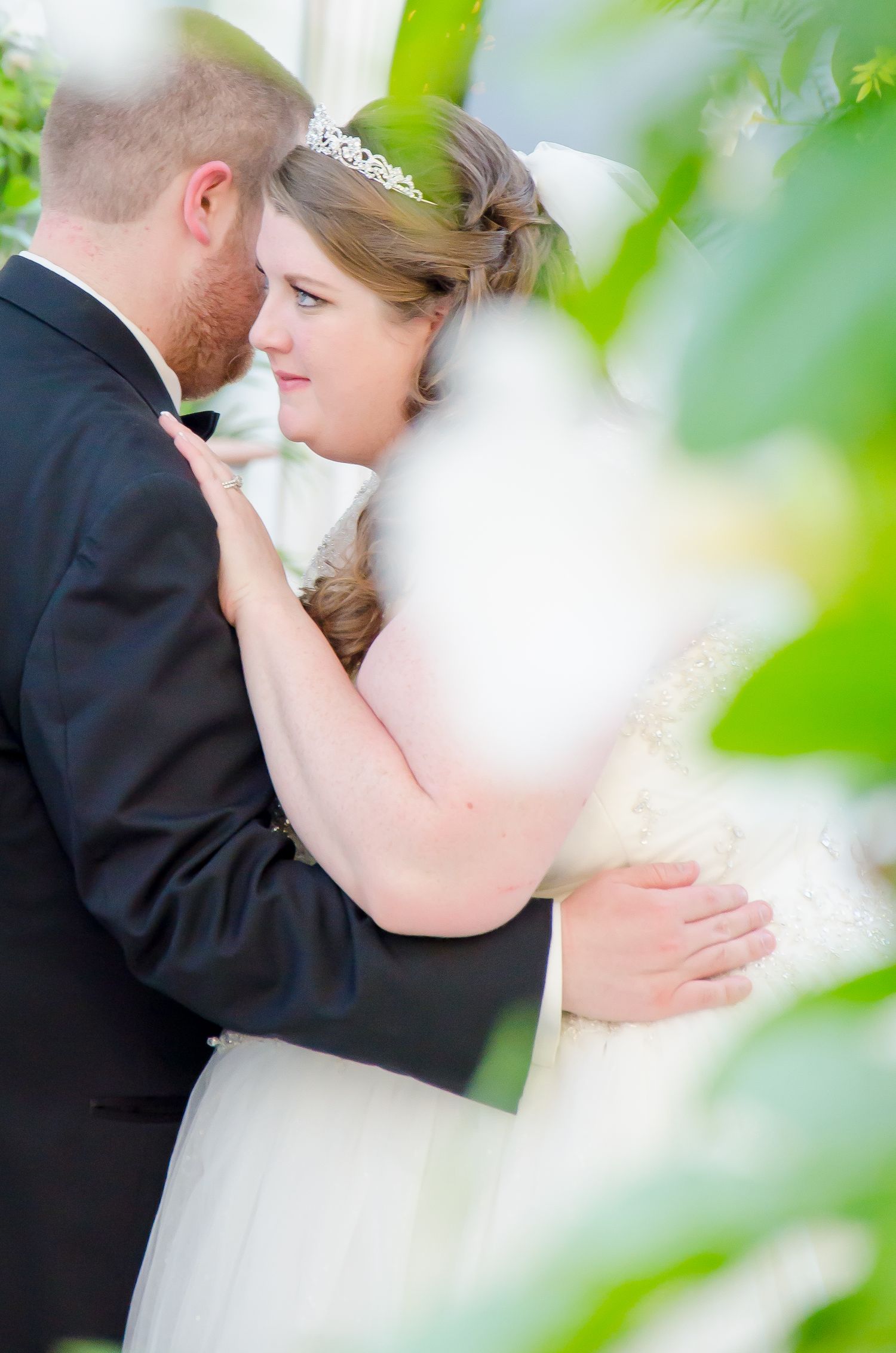 A bride and groom share a romantic embrace outdoors, framed by lush green leaves, as she wears a tiara and white gown.