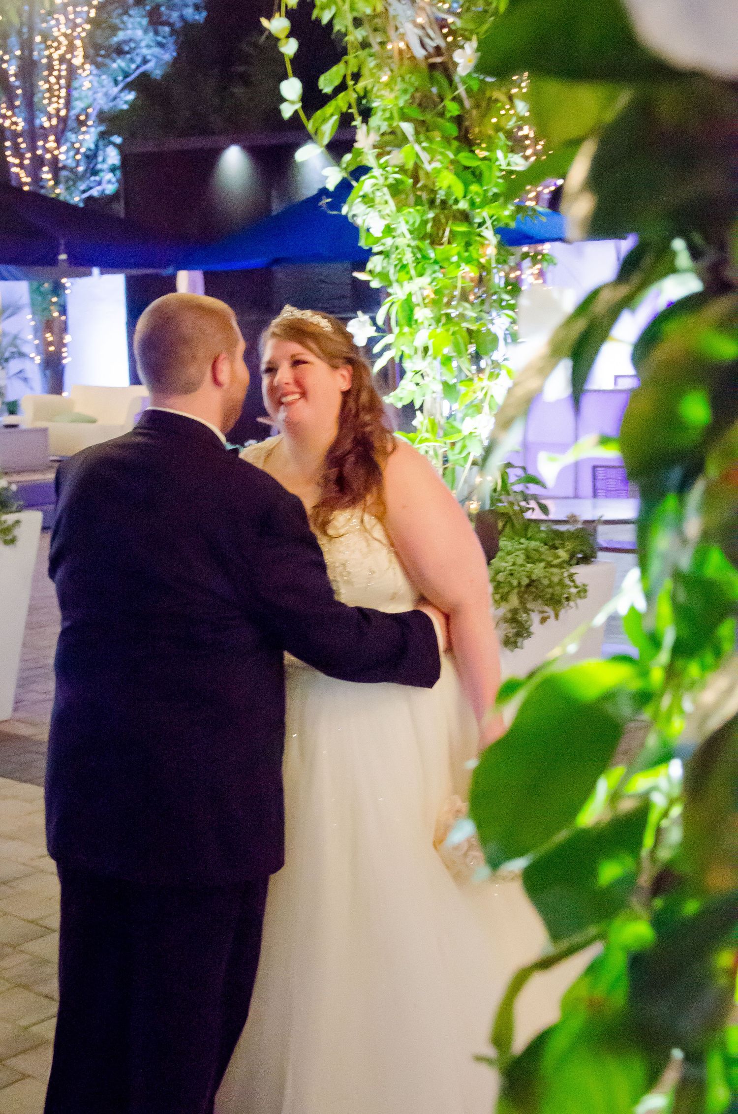 Smiling bride in white gown shares romantic moment with groom surrounded by lush greenery at wedding.