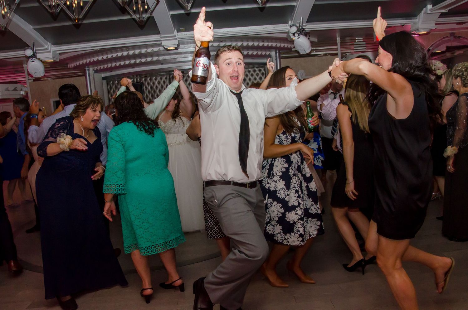 Man dancing joyfully with champagne bottle at lively wedding reception party with guests on dance floor.