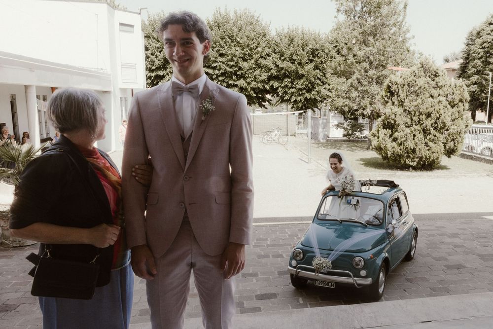 Groom in beige suit stands with elderly woman outdoors, vintage blue Fiat 500 with passenger visible in background.