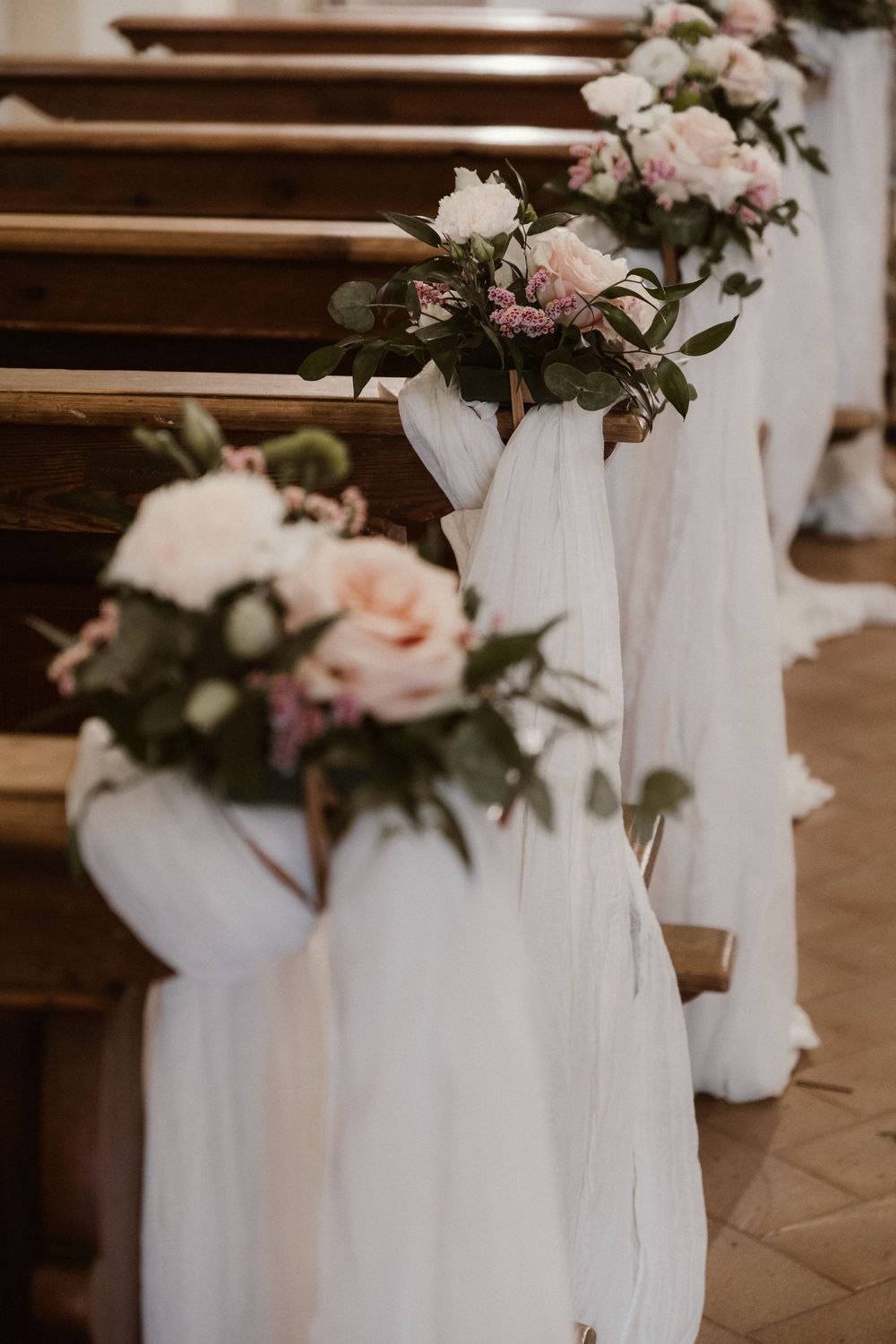 Church pews decorated with white fabric draping and blush pink floral bouquets for an elegant wedding ceremony.