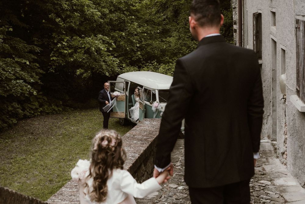 Groom holds flower girl's hand walking toward vintage wedding car at outdoor venue.