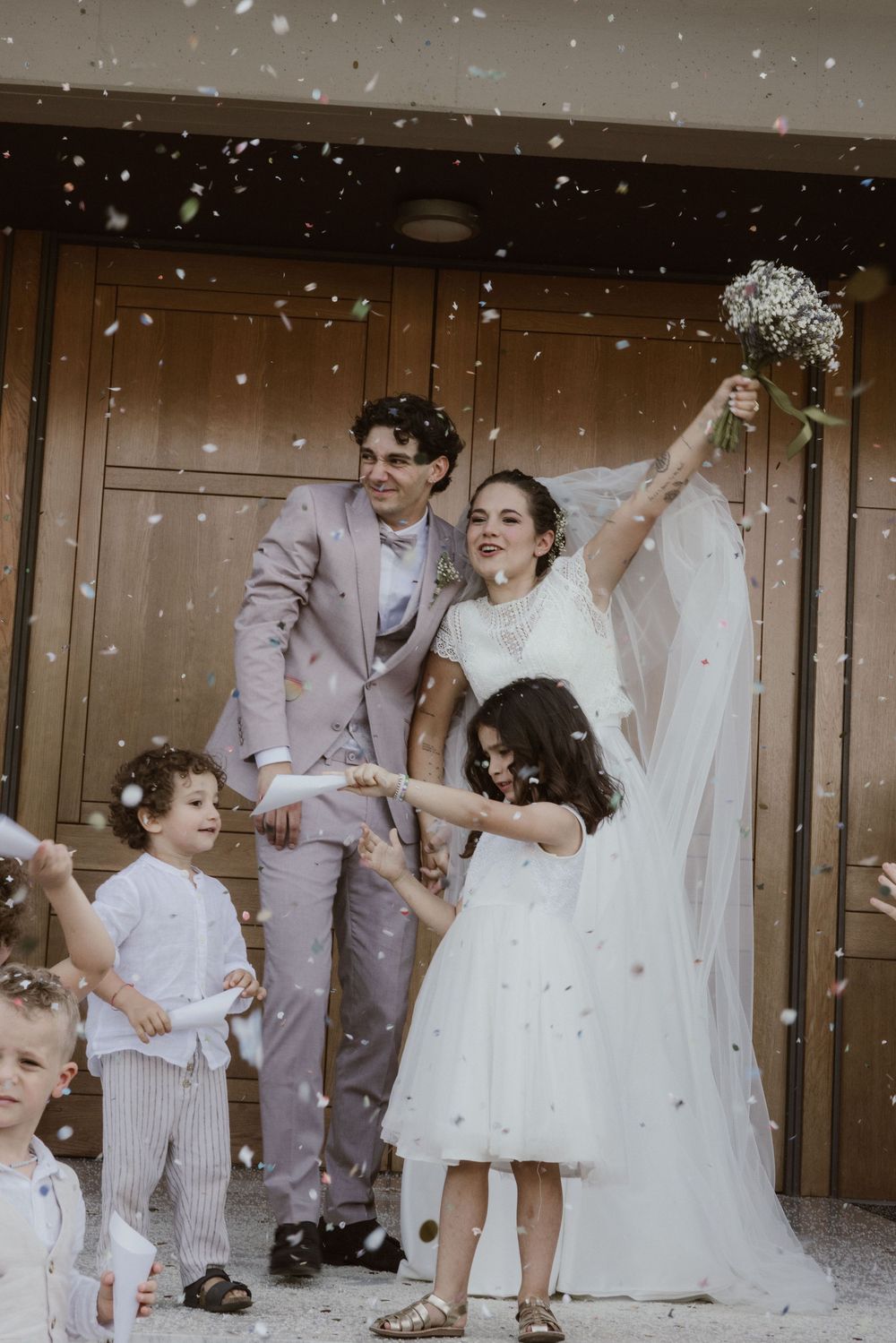 Joyful newlyweds celebrate with confetti as bride raises bouquet, flanked by children in wedding attire outside church doors.
