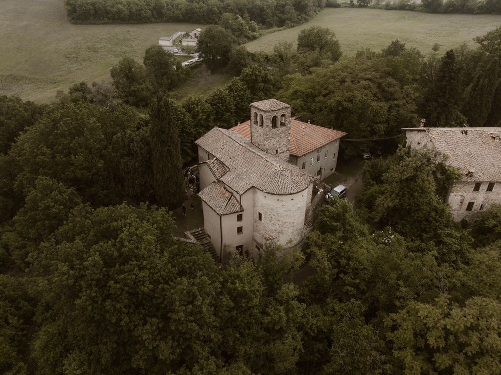 Aerial view of an ancient stone church surrounded by dense green trees in the Italian countryside.