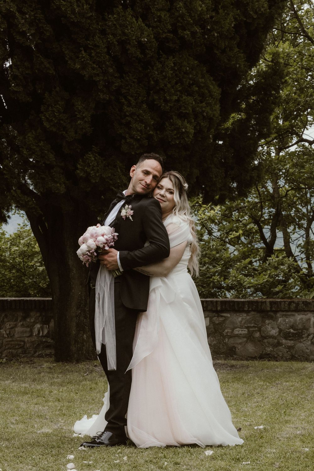 Bride and groom embrace outdoors in wedding attire, holding pink bouquet, with lush green trees in background.