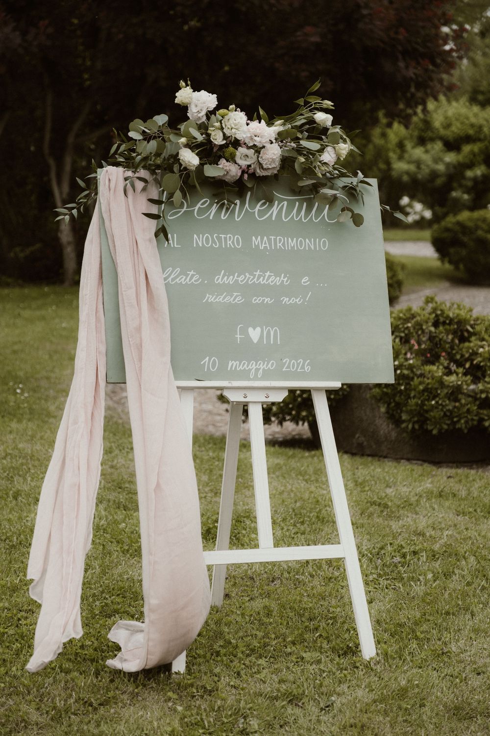 Elegant wedding welcome sign on white easel with floral arrangement and blush ribbon, displayed outdoors on green lawn.
