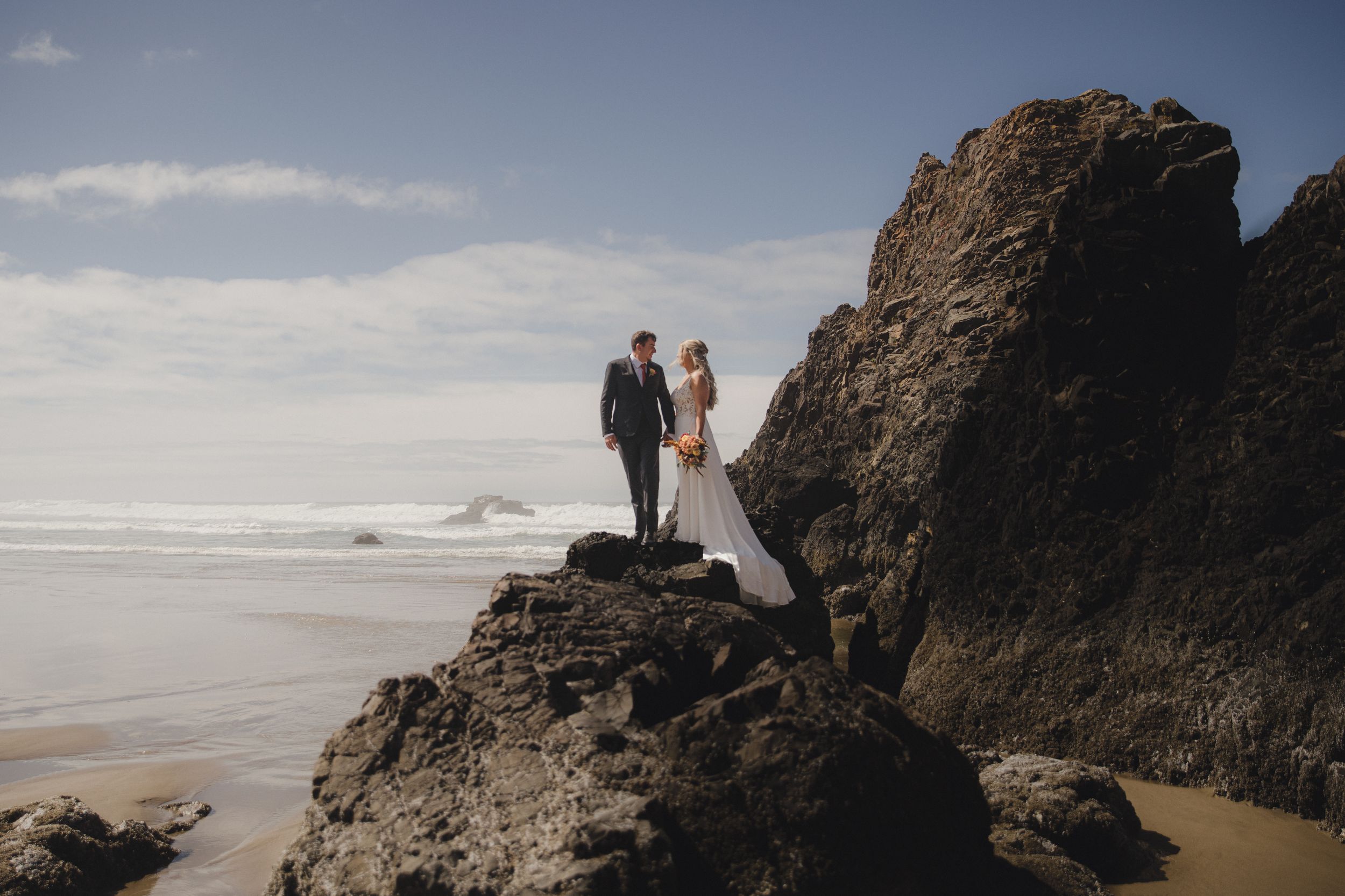 Bride and groom standing on rocky coastal cliffs with ocean waves and blue sky backdrop at beach wedding.