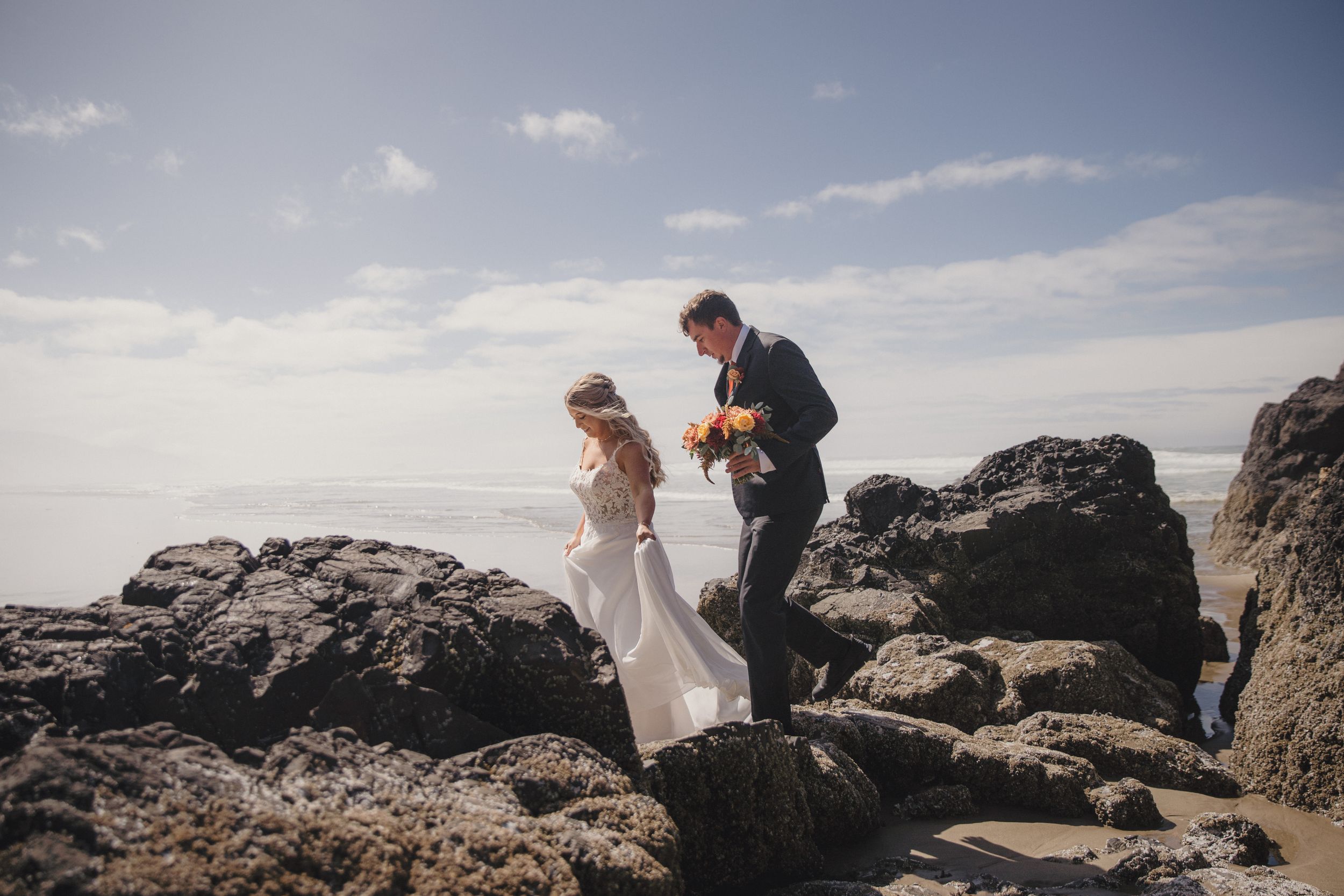 Bride and groom carefully navigating rocky coastal terrain during their outdoor beach wedding photo session.
