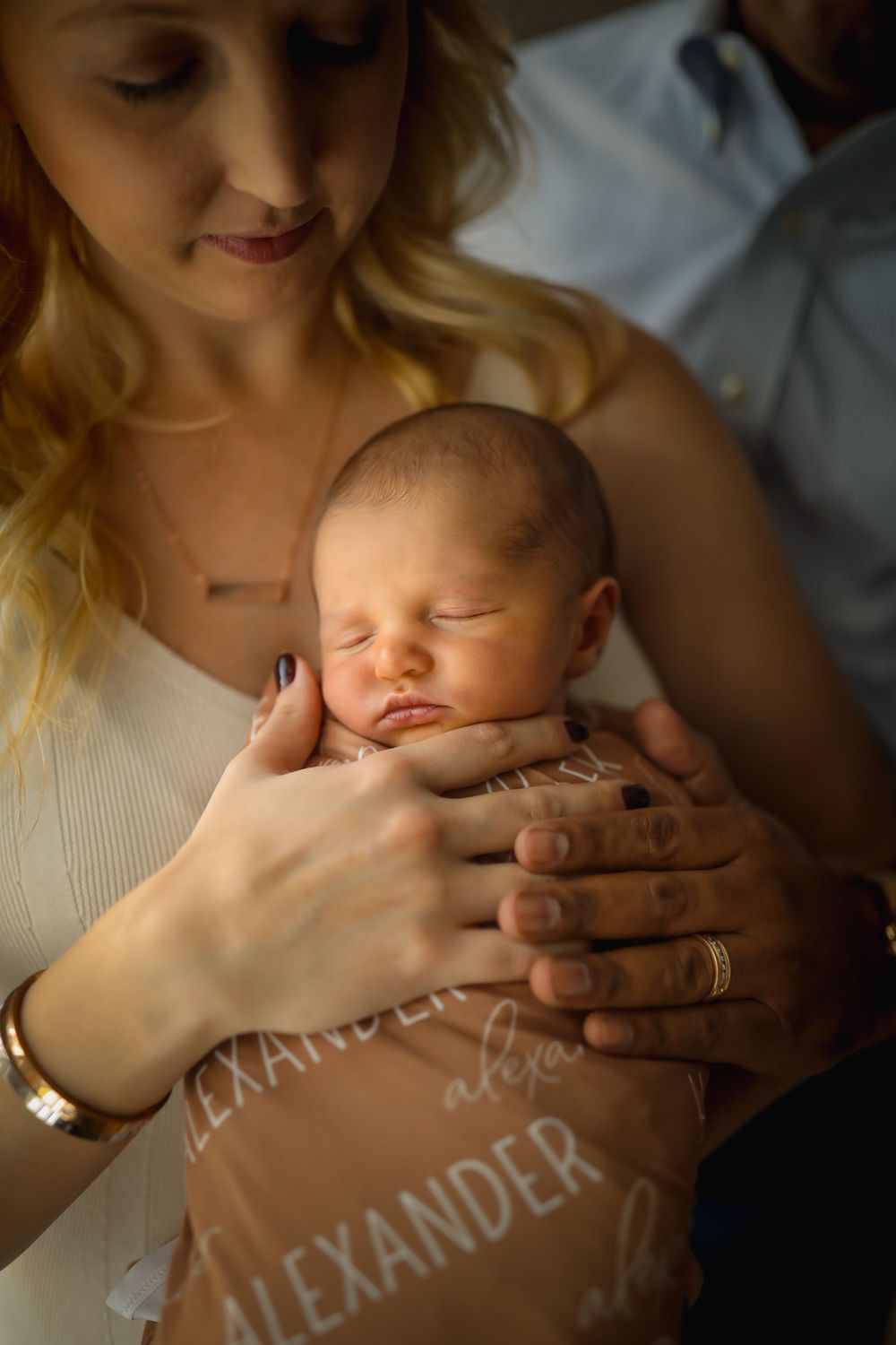 Parents tenderly cradling sleeping newborn baby Alexander, wrapped in personalized name blanket.