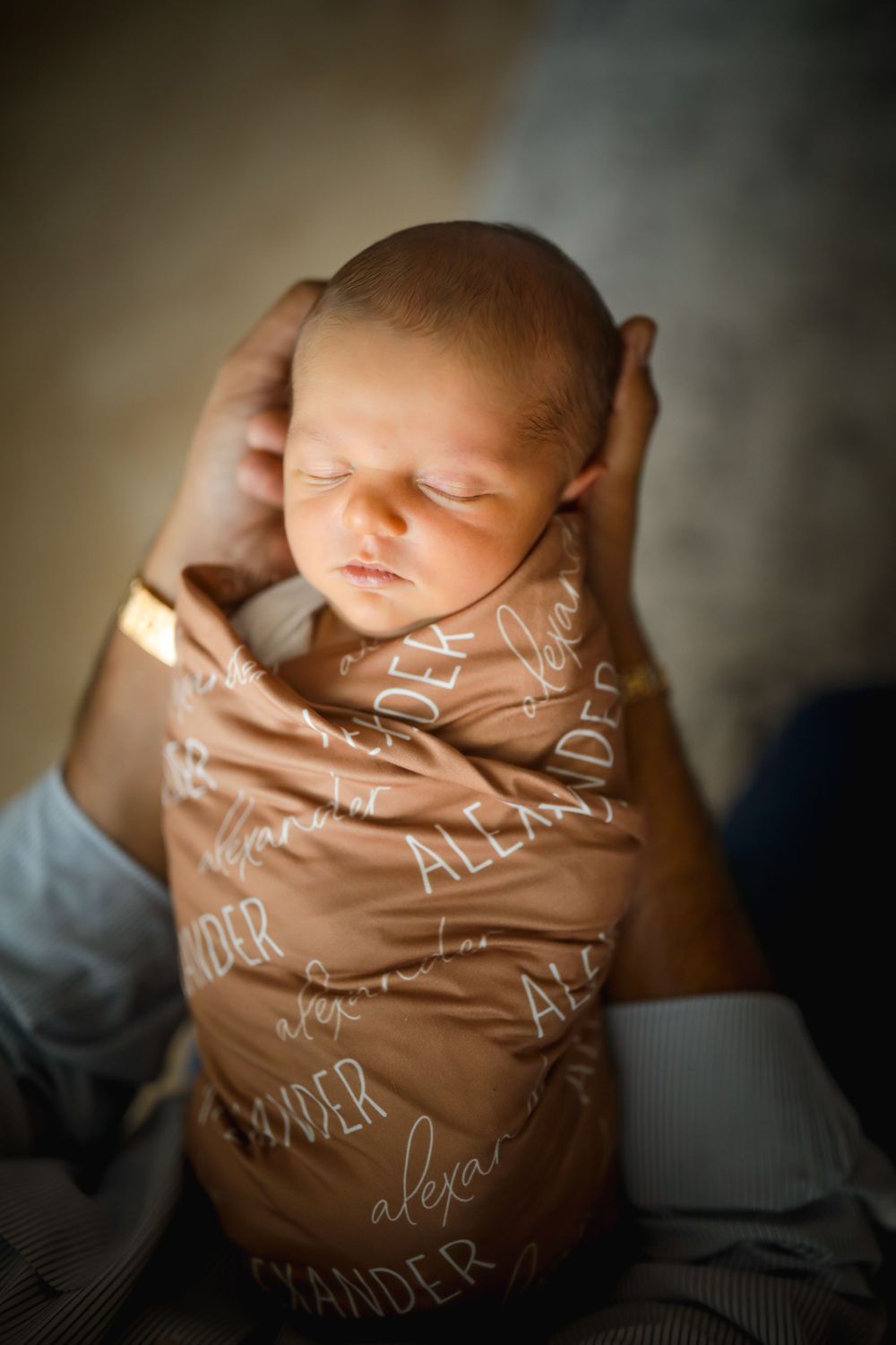Sleeping newborn baby swaddled in a personalized brown blanket with the name Alexander, cradled in adult hands.