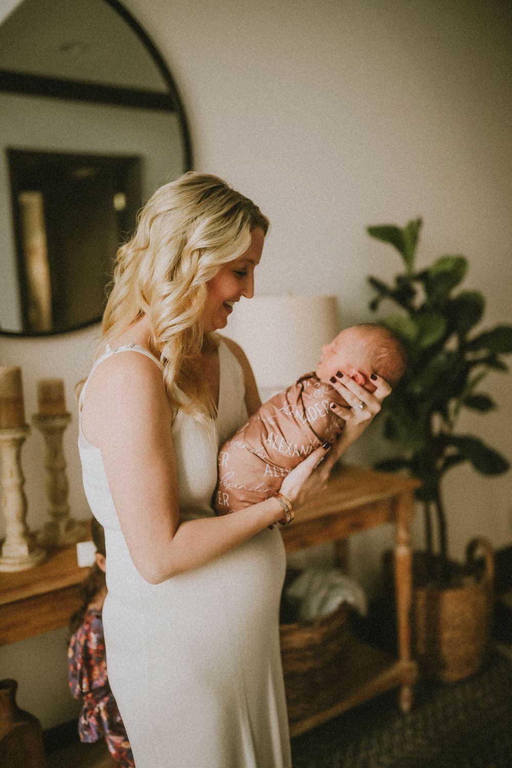 Blonde mother in white dress tenderly holding swaddled newborn baby in cozy home setting.