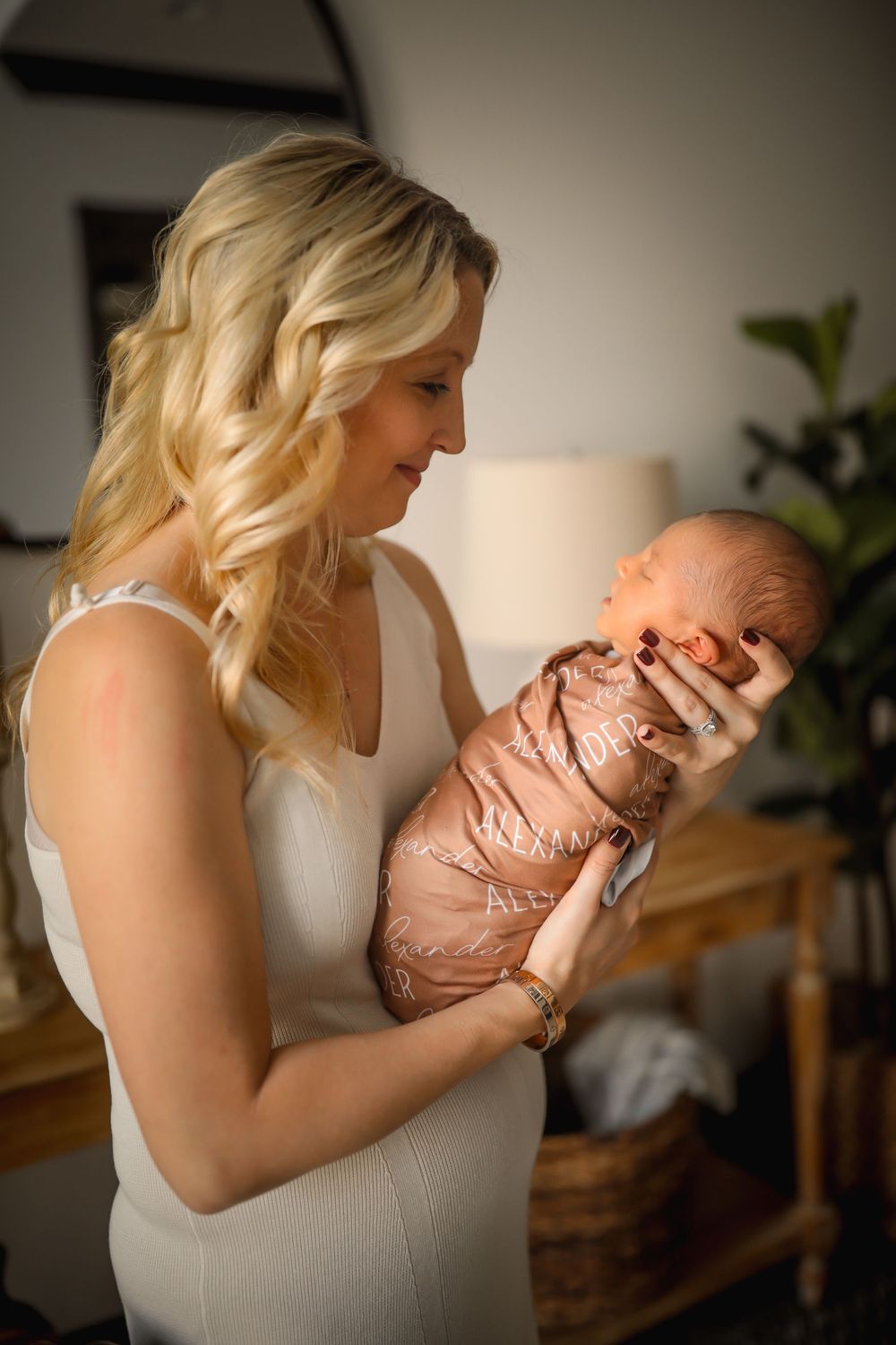 A blonde woman in a white dress tenderly holds a swaddled newborn baby indoors.