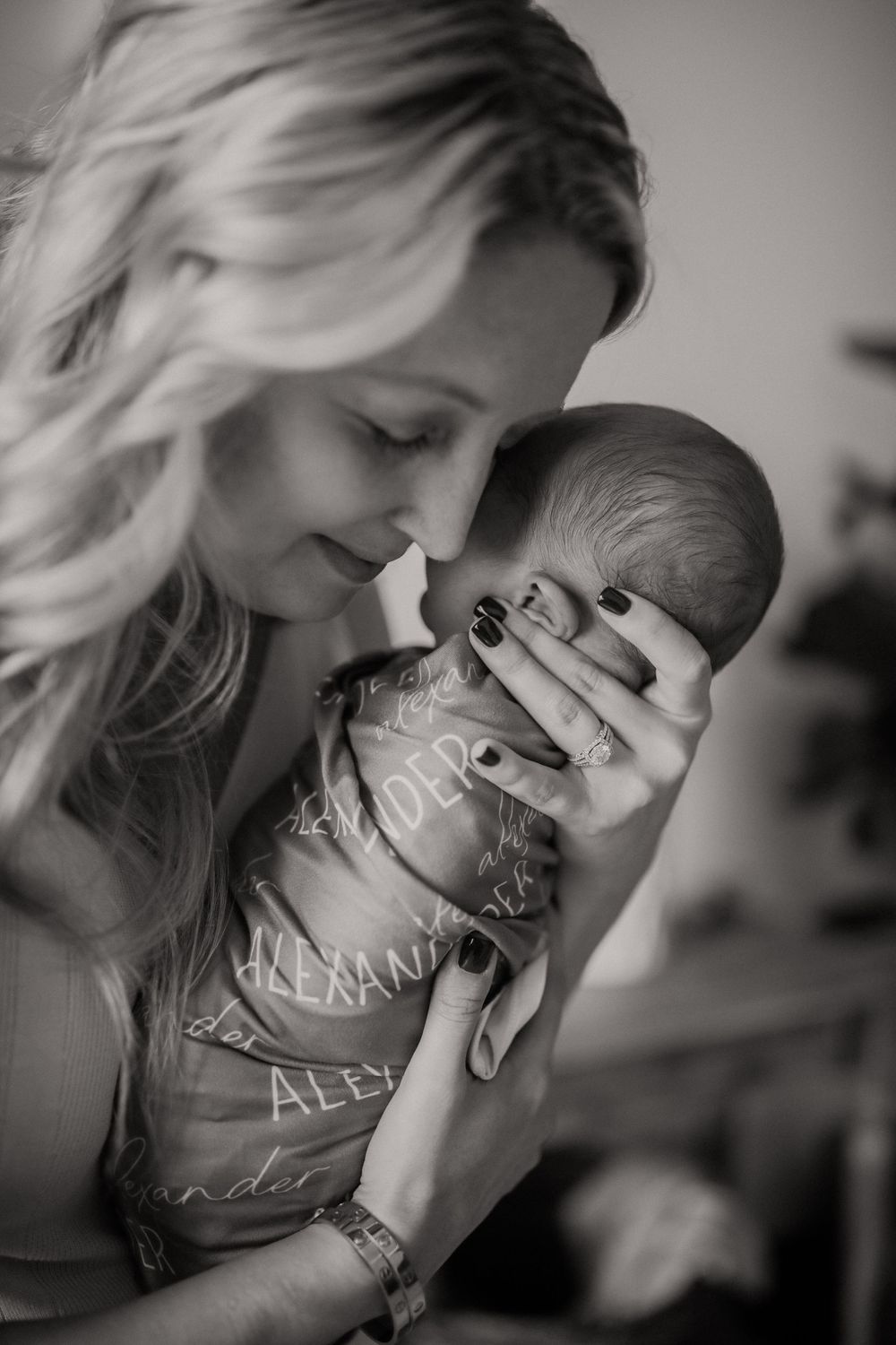 Black and white photo of a blonde mother tenderly holding and nuzzling her newborn baby wrapped in a personalized swaddle.