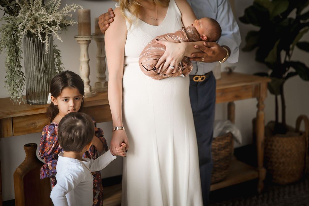 Family portrait with mother in white dress holding newborn, father nearby, and two young children standing close.