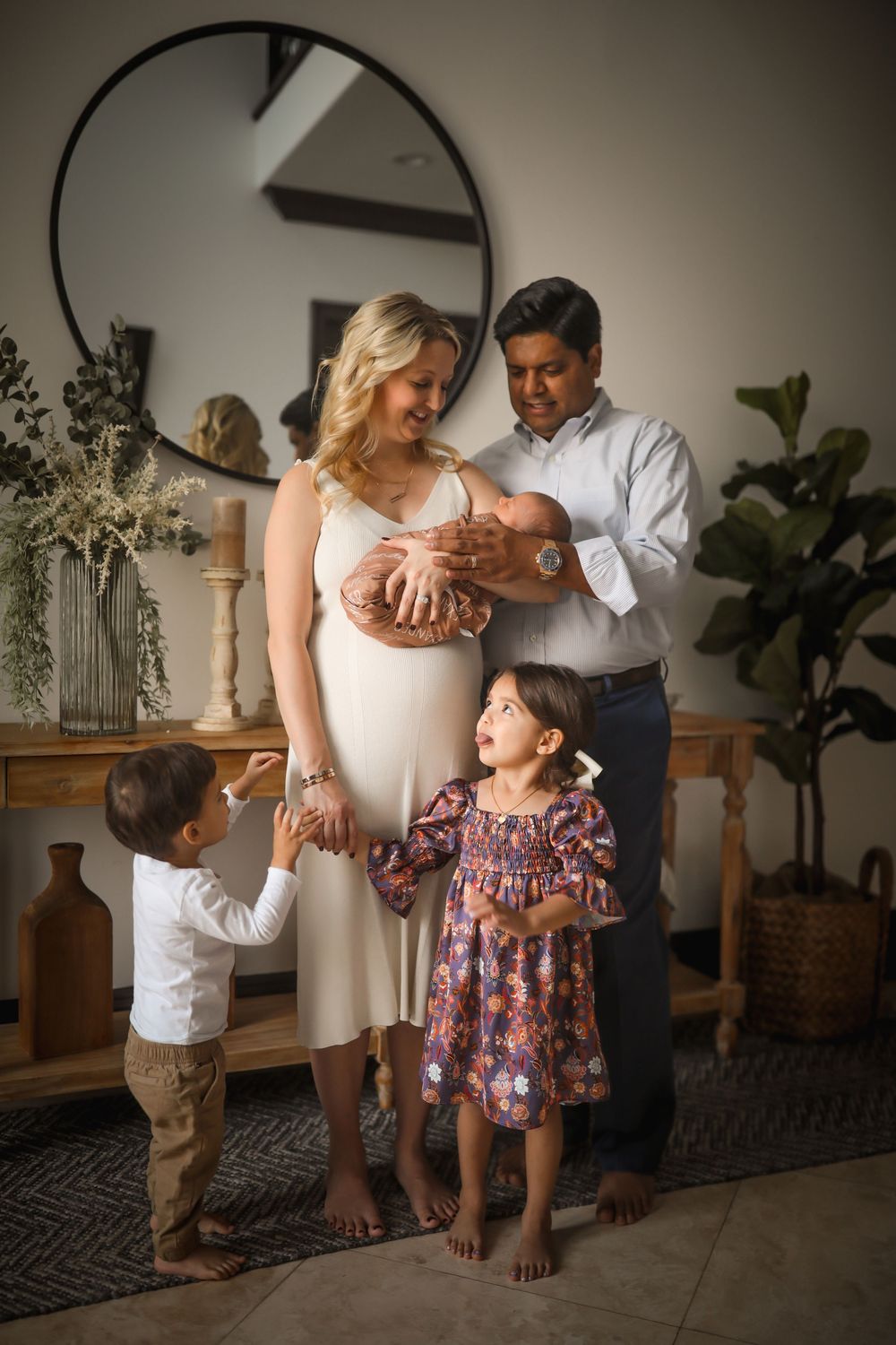 Family portrait with parents holding newborn baby while two young children stand nearby in a bright home interior.