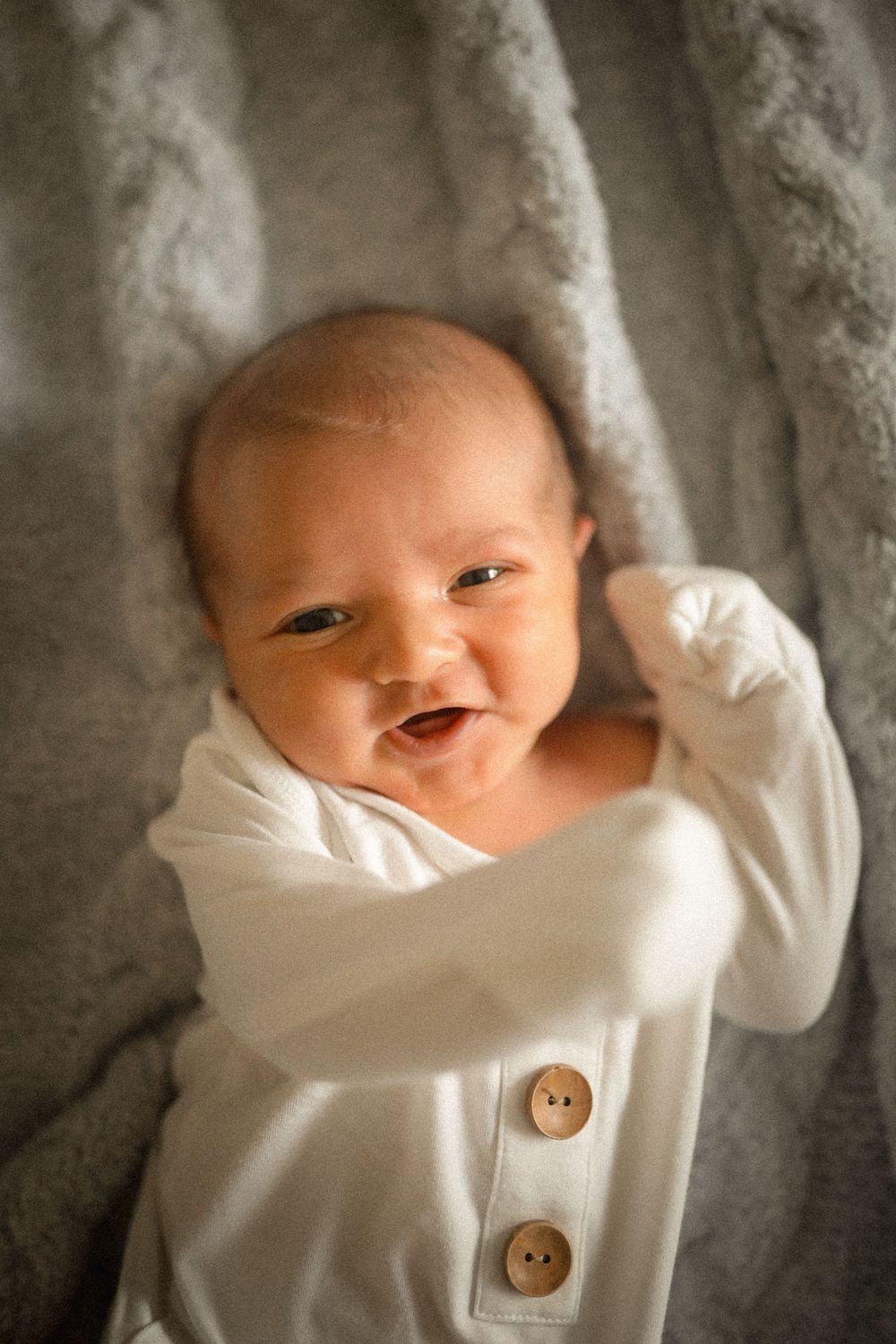Smiling newborn baby in white button onesie lying on soft gray knit blanket.