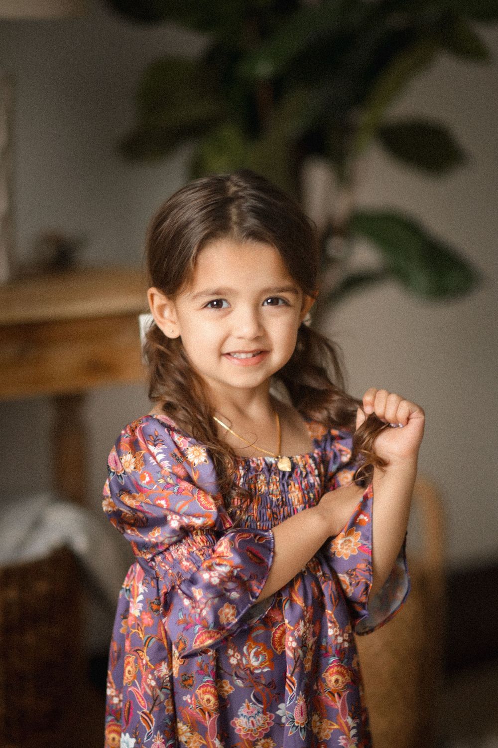 A smiling young girl in a floral purple dress holds her braided hair while posing indoors.