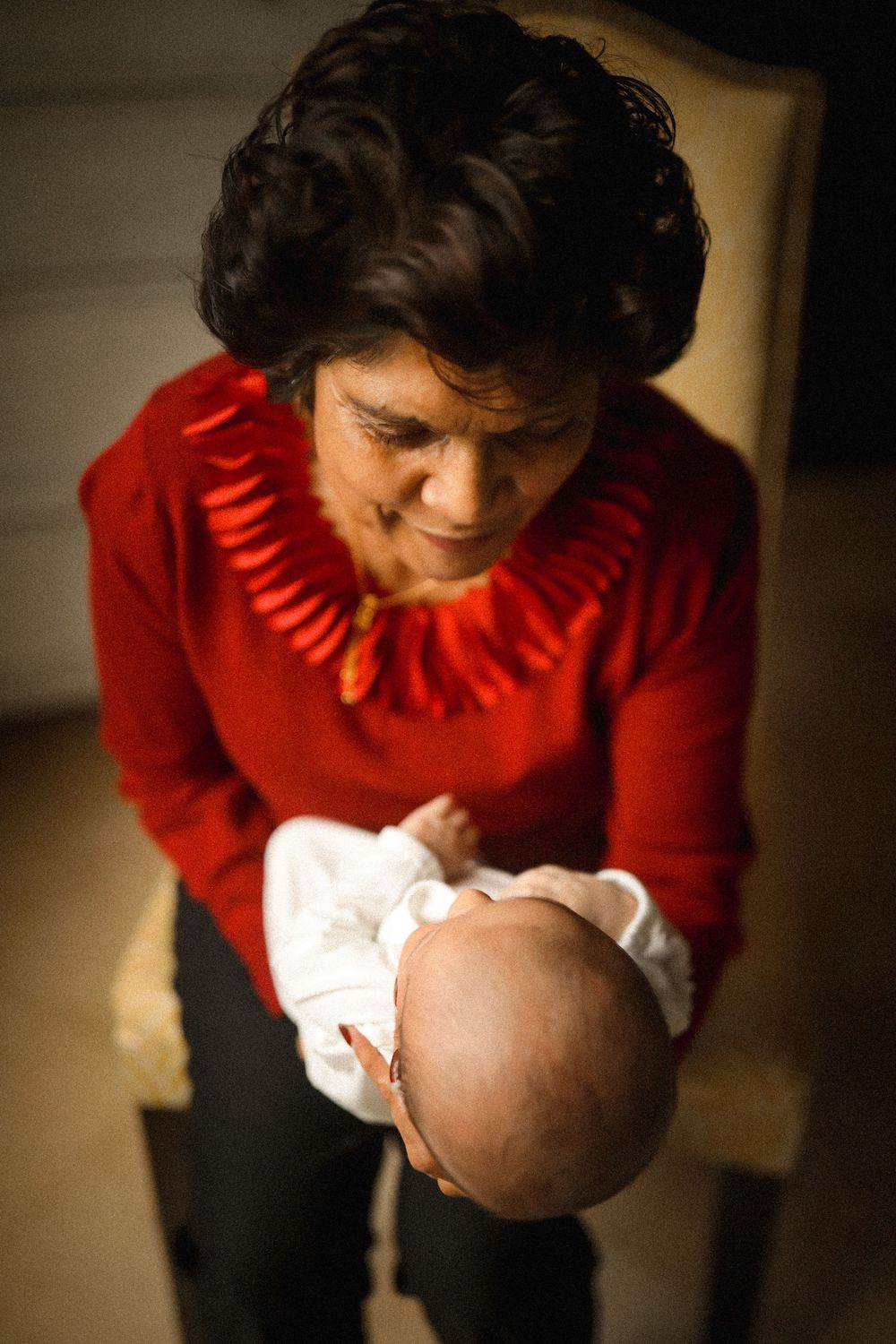 Grandmother in red sweater tenderly holds sleeping newborn baby while seated in a chair.