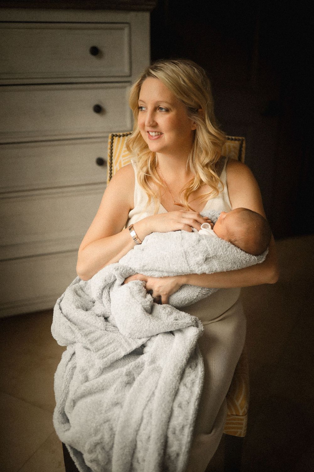 A smiling blonde mother sits in a chair cradling her newborn baby wrapped in a soft gray blanket.