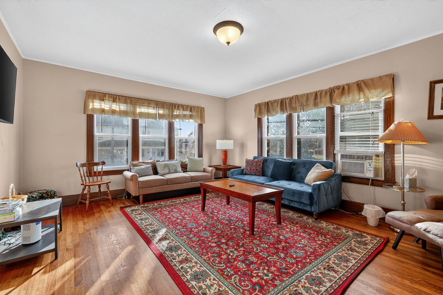 Bright living room with hardwood floors, blue couch, red area rug, crown molding, and large windows.