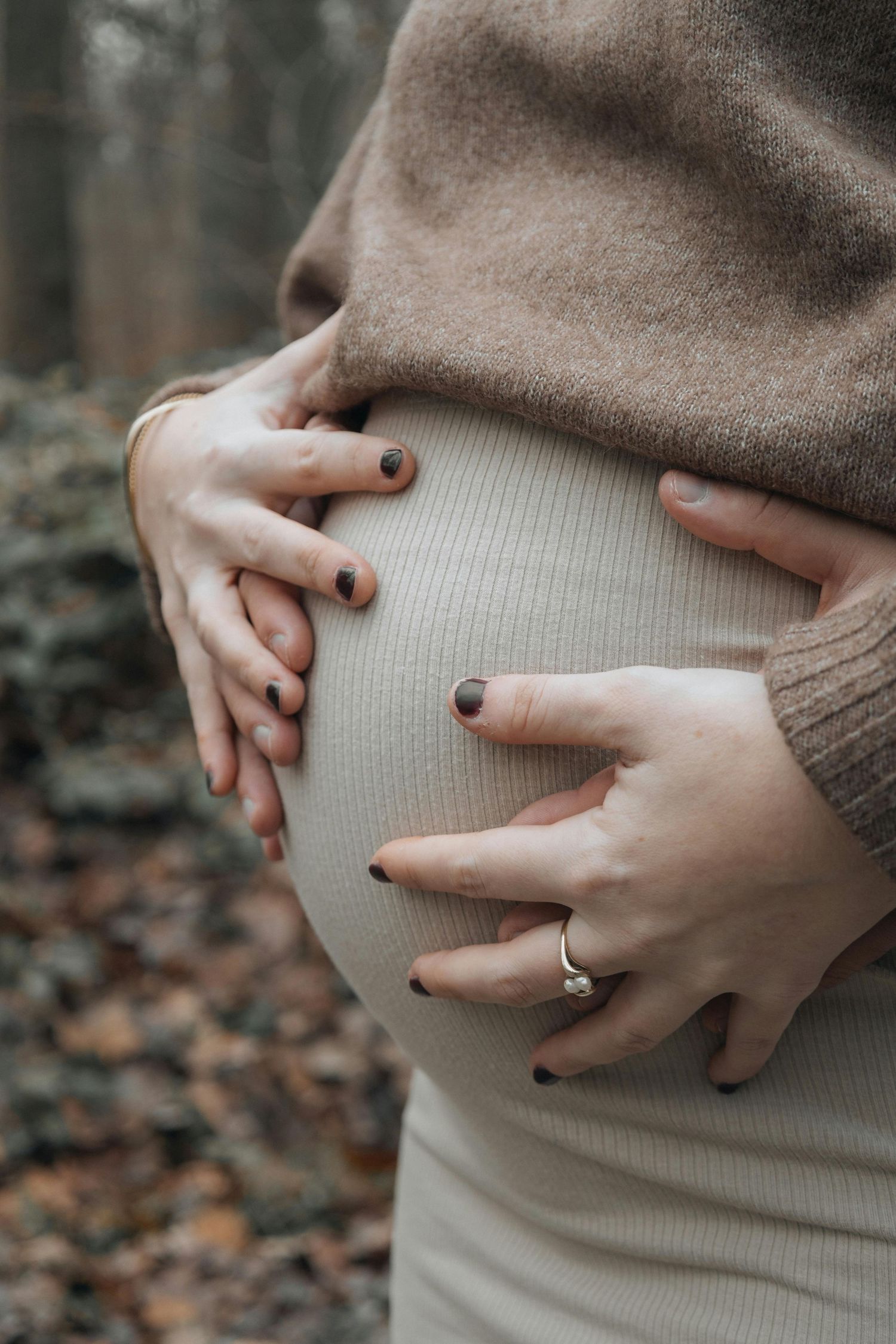 Femme enceinte vêtue de beige, caressant son ventre rond à deux mains dans une forêt d'automne