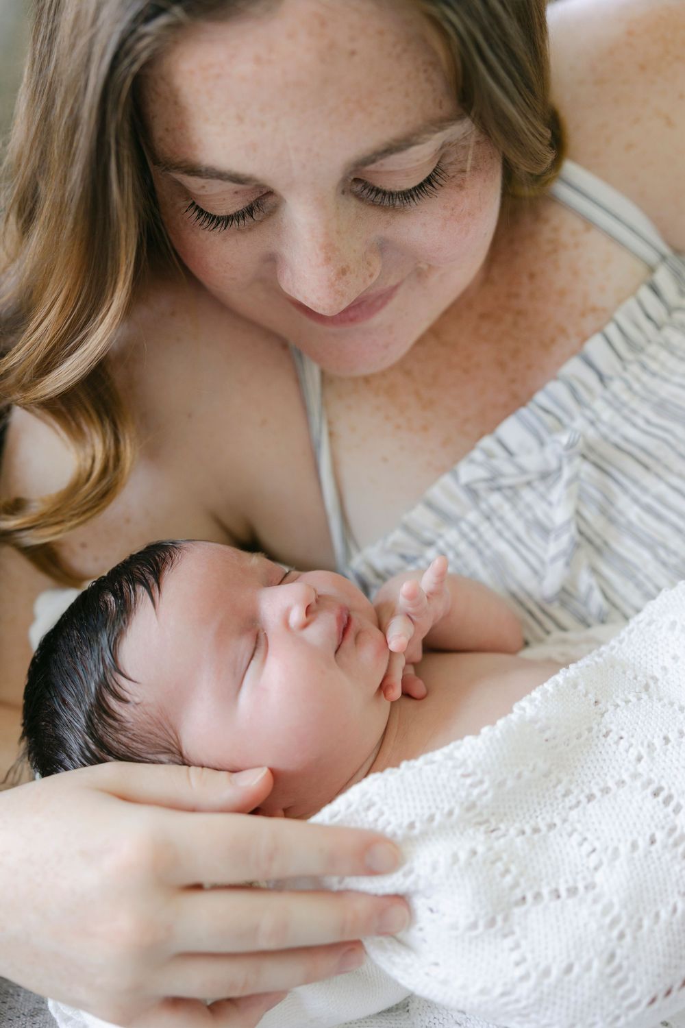 A freckled mother with auburn hair smiles down at her sleeping newborn baby wrapped in a white knit blanket.