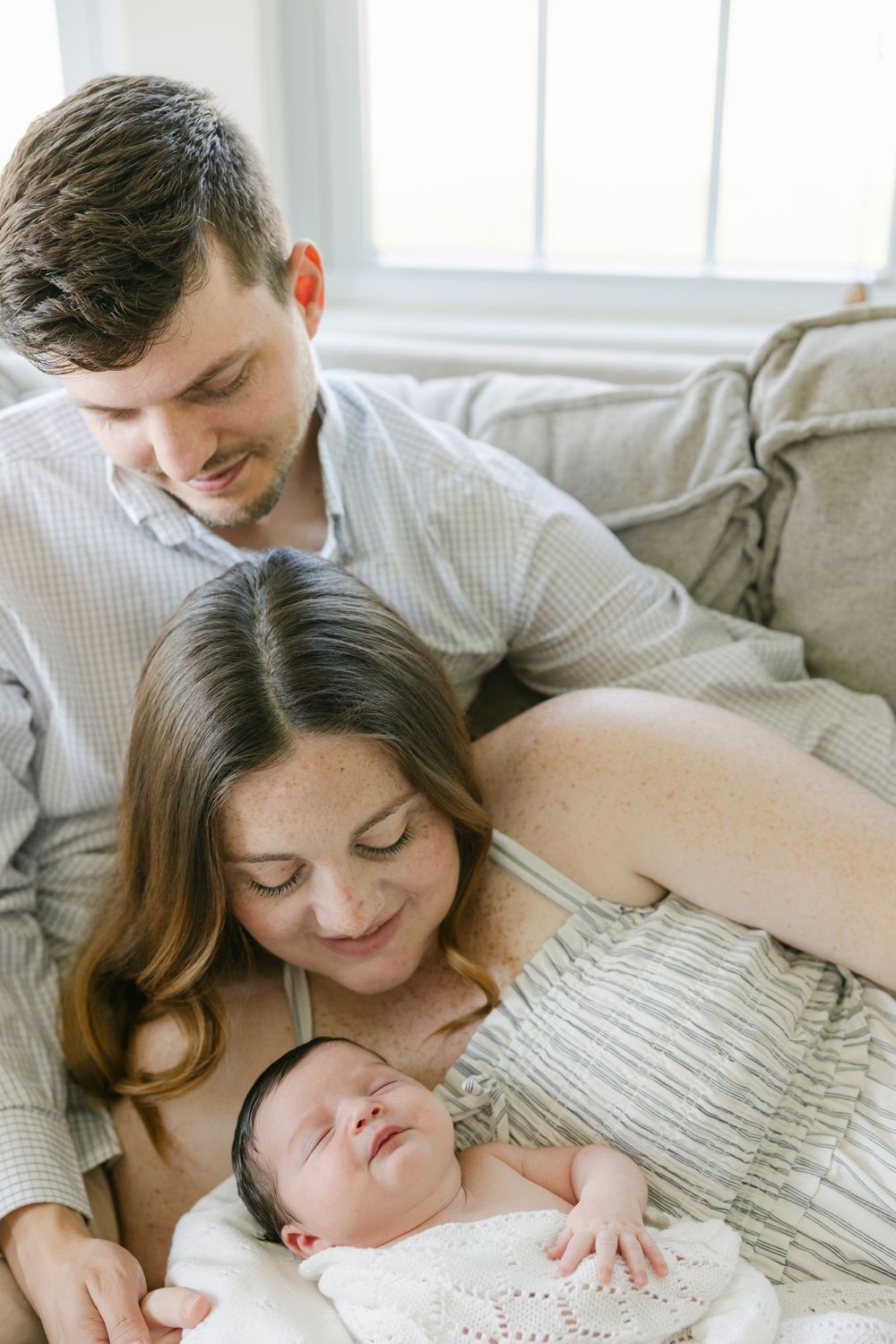 New parents lovingly gaze at their sleeping newborn baby wrapped in white lace blanket on a cozy sofa.