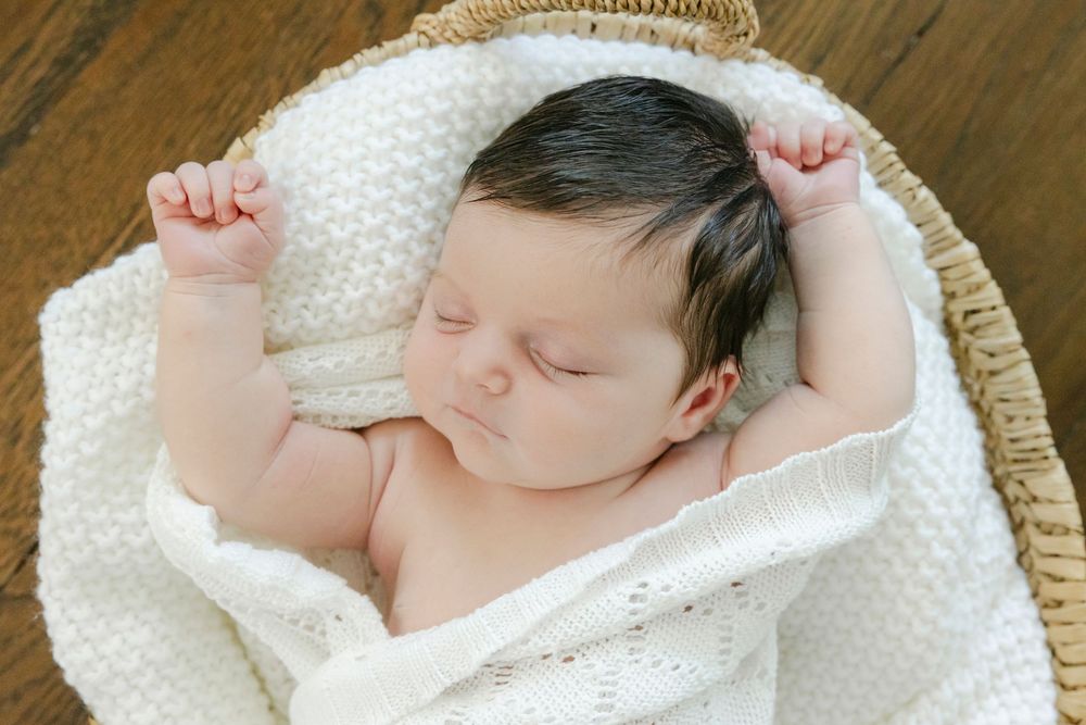 Peaceful newborn baby sleeping in a wicker basket wrapped in a soft white knit blanket with tiny fists raised.