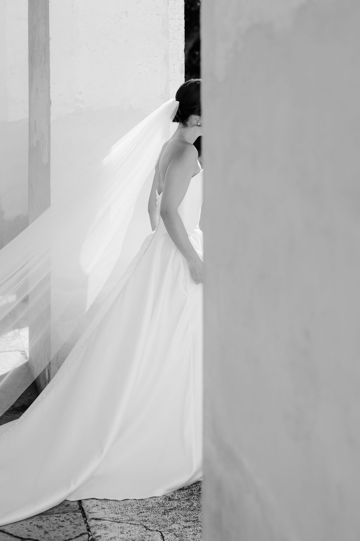 Black and white photo of a bride in a flowing white gown and long veil standing between white walls.