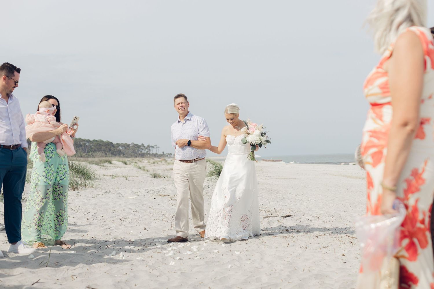 Bride and groom celebrate on a sandy beach wedding ceremony with guests cheering in background on a clear day.