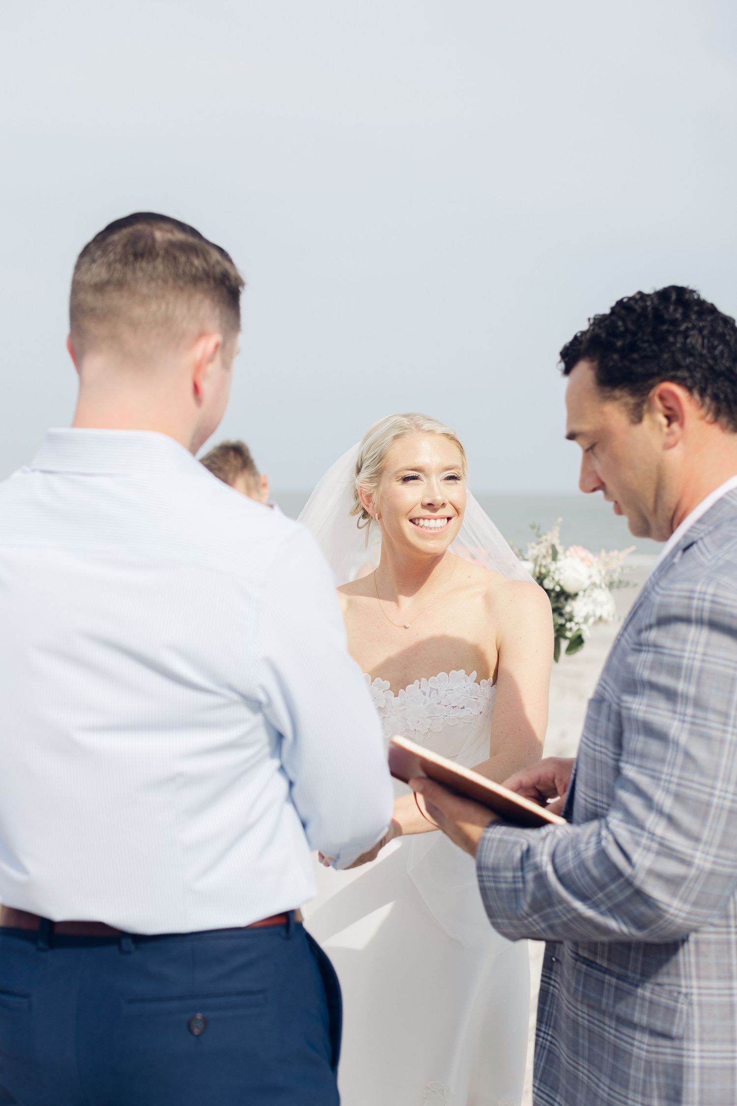 Smiling bride in white gown and veil exchanges vows with groom during outdoor beach wedding ceremony.