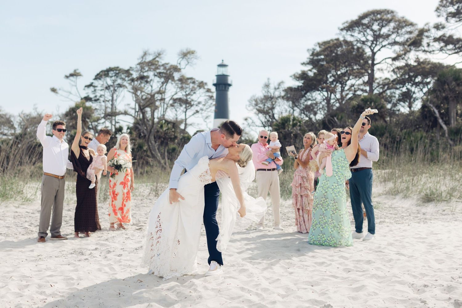 Couple shares romantic dip kiss on sandy beach with wedding guests celebrating near a lighthouse backdrop.
