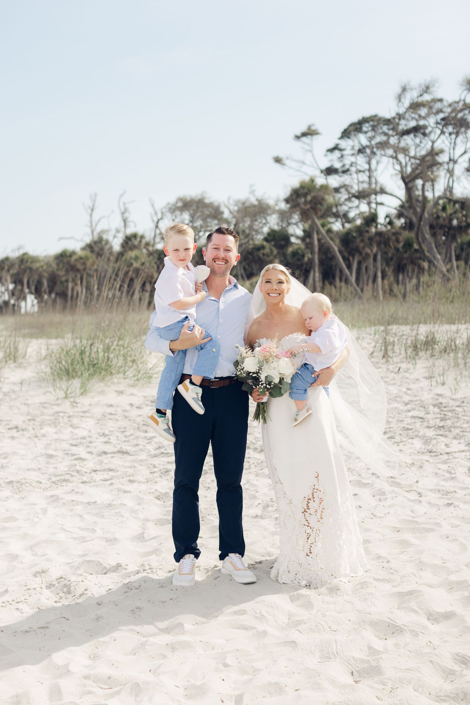 Bride and groom with two young children pose on sandy beach, bride holding bouquet in lace wedding gown.