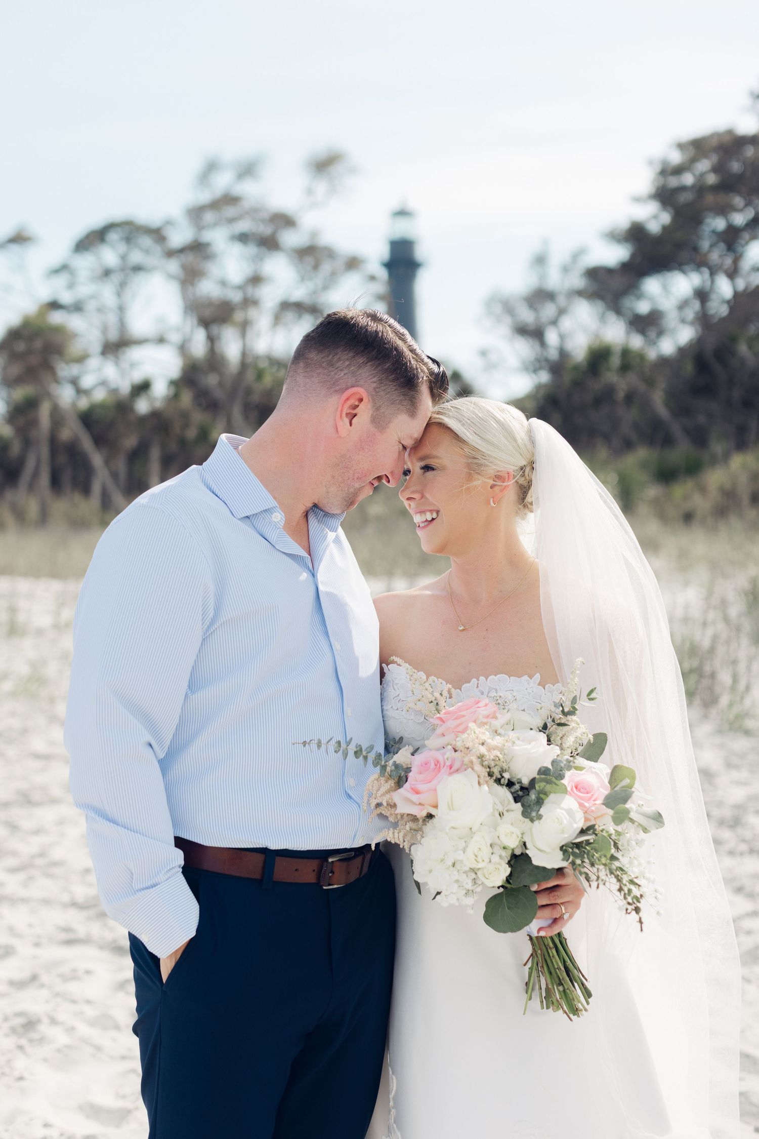 Bride and groom share a romantic beach moment, bride holding pink and white bouquet with lighthouse in background.
